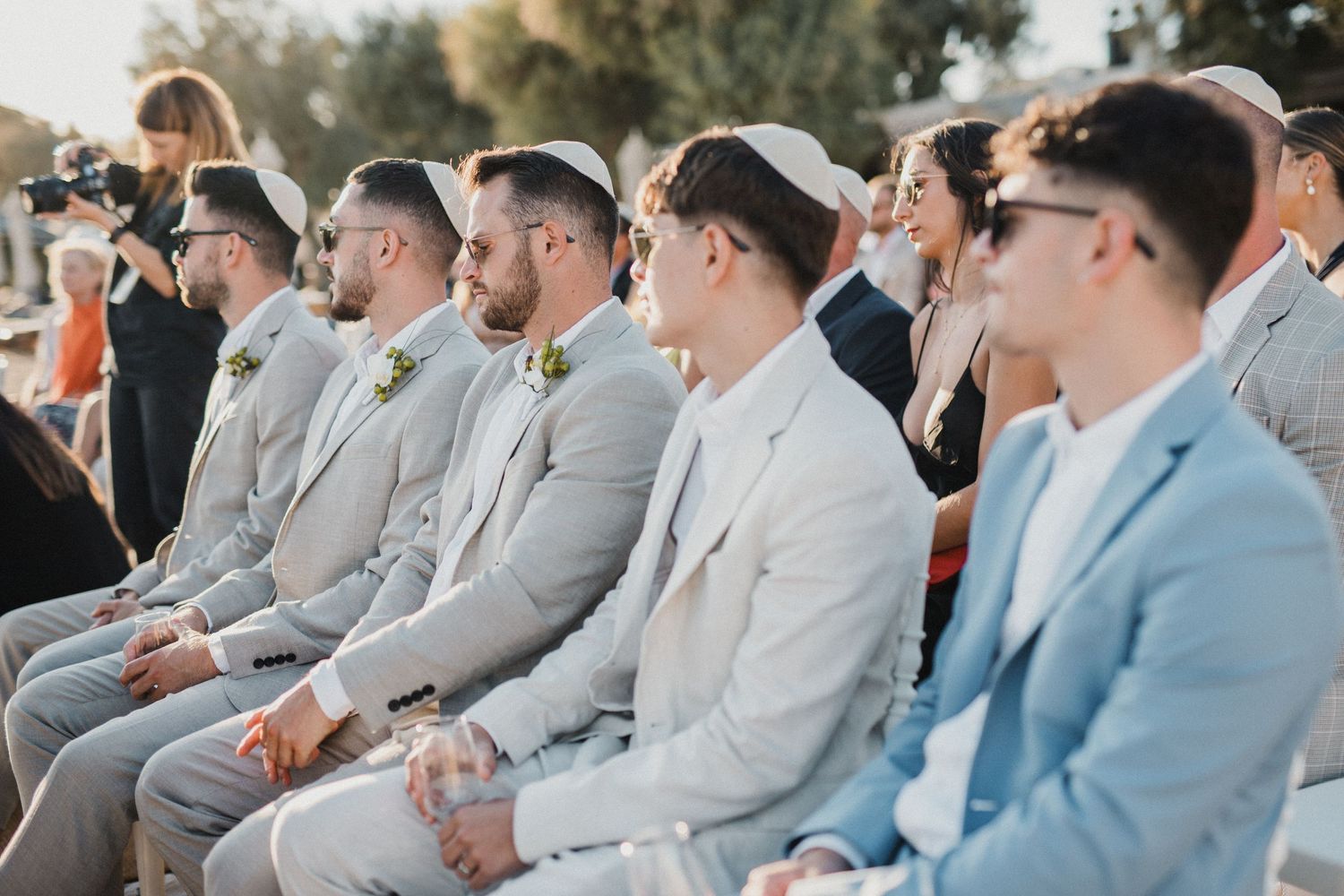 Wedding guests in light colored suits seated at an outdoor ceremony.
