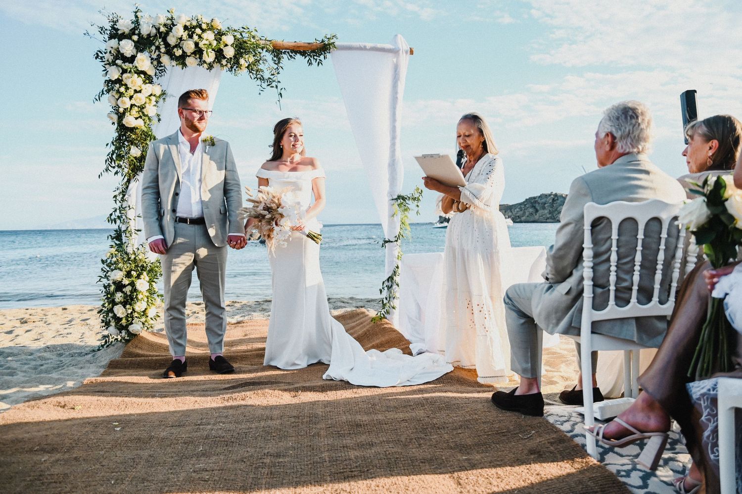 Beach wedding ceremony with floral arch and white chairs overlooking ocean.