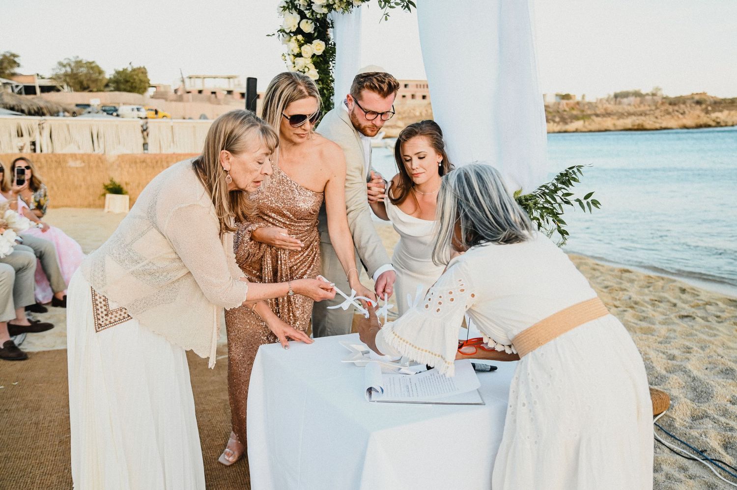 Wedding guests gather around a white-clothed table on a beach to sign the guest book during a seaside ceremony.