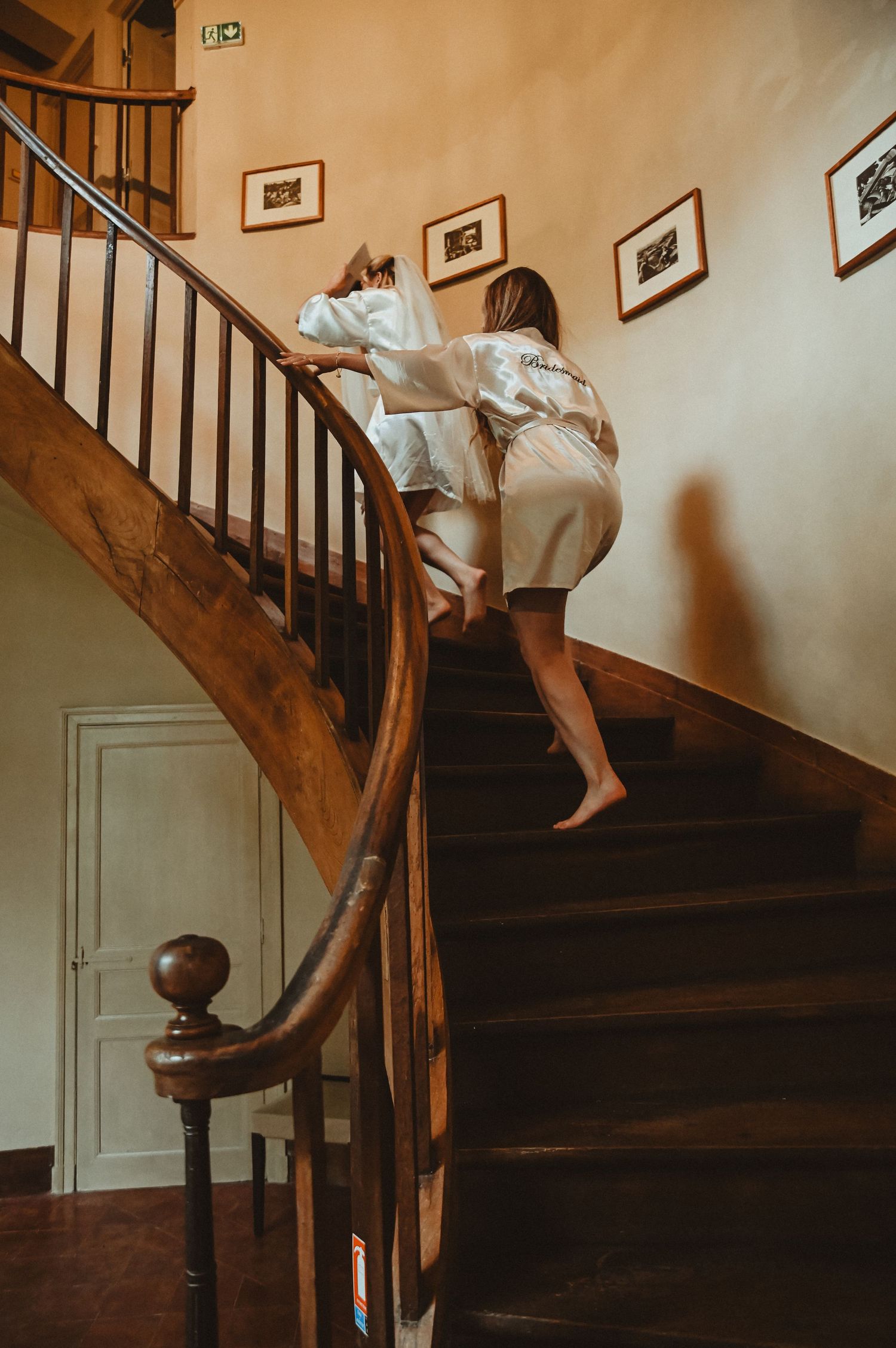 Two people in white robes climbing an elegant wooden staircase with framed artwork on the wall.
