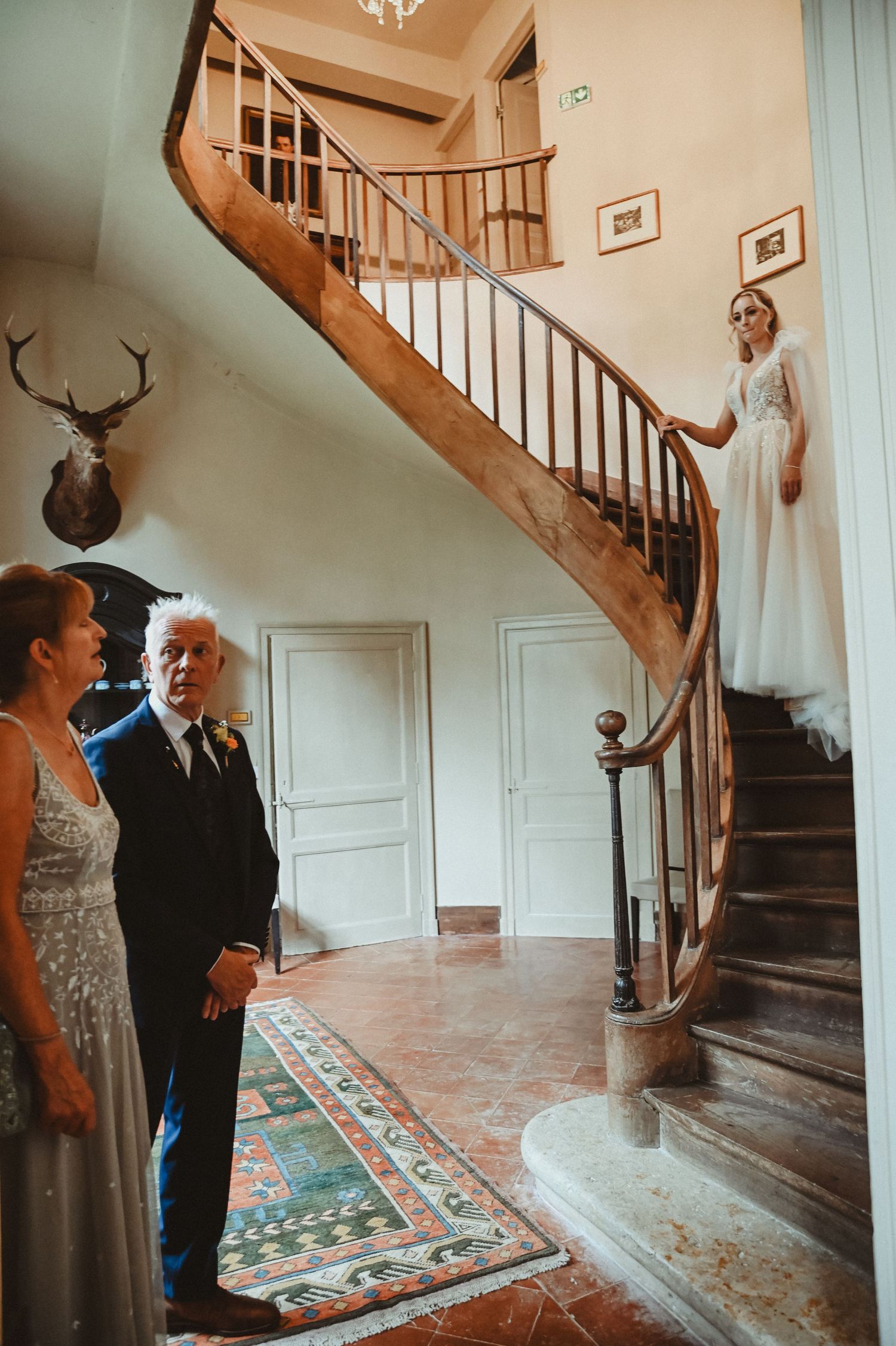 A vintage staircase decorated with a mounted deer head features an elegant curved wooden banister and patterned rug.