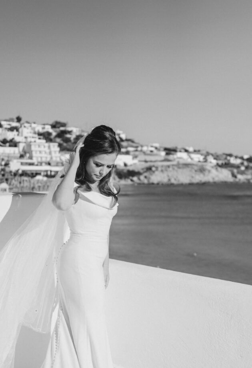 Classic white wedding gown silhouette photographed against a coastal Mediterranean backdrop in black and white.