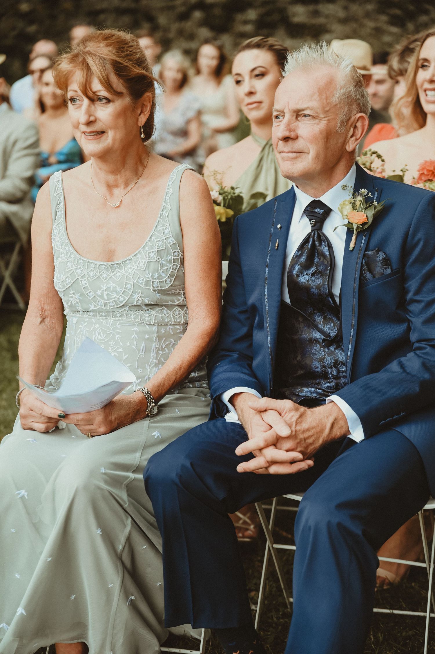 Wedding guests in formal attire watch the ceremony from folding chairs in a garden setting.