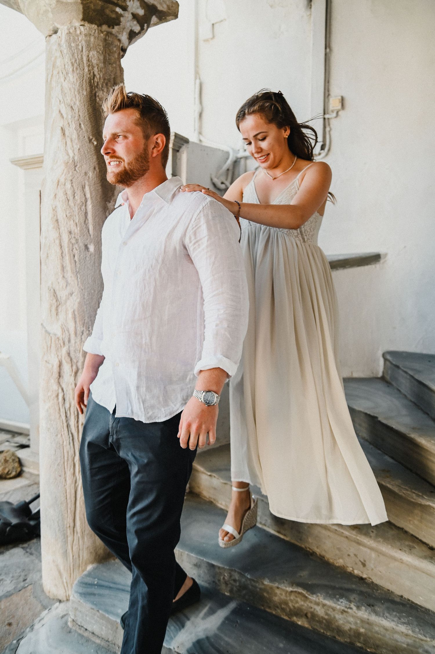 Couple in white clothing posing playfully on stone steps against whitewashed Mediterranean architecture.
