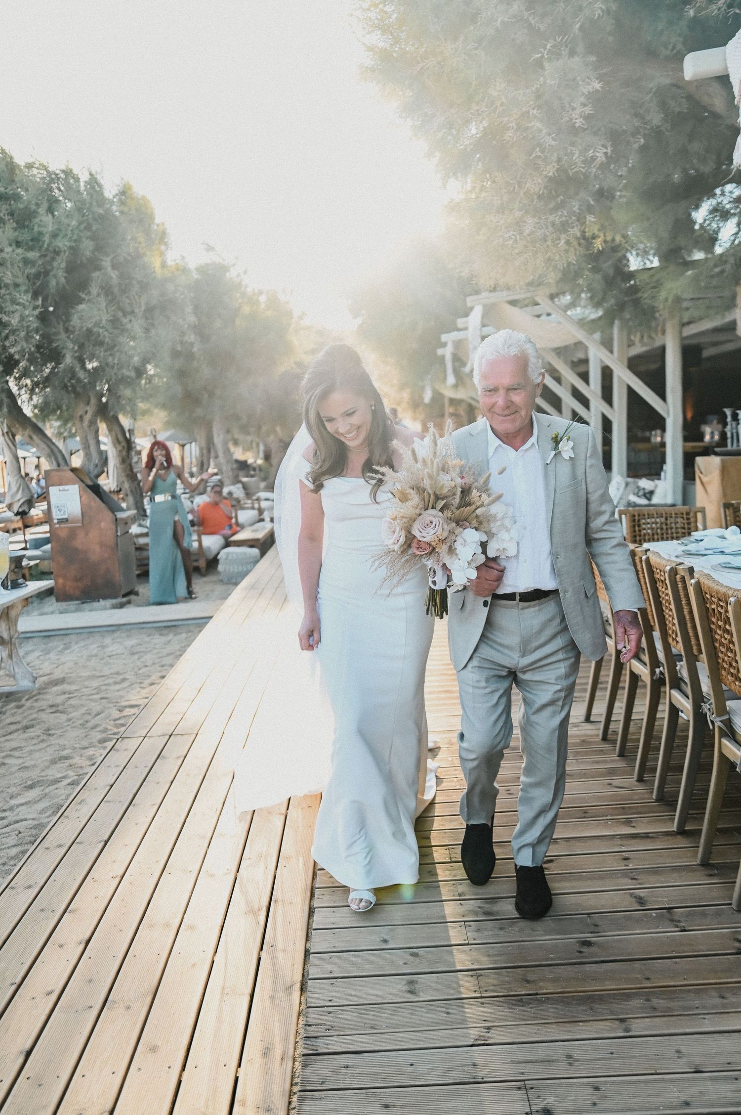Sunlit outdoor wedding procession on a wooden boardwalk with elegant floral arrangements.