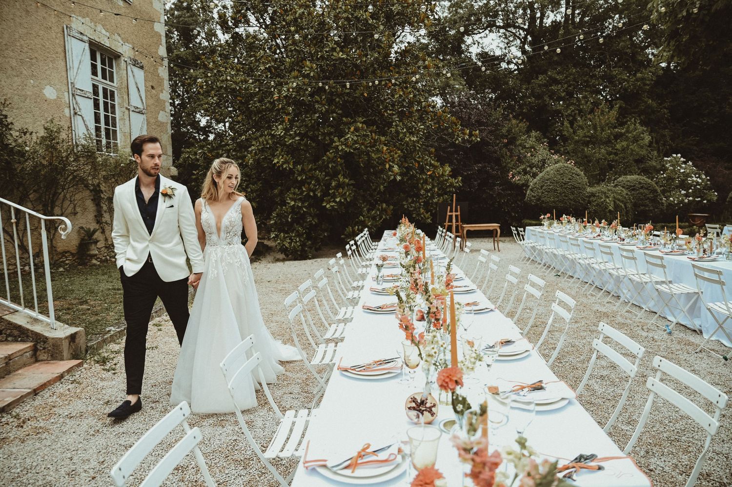 Outdoor wedding reception setup with long tables decorated with floral centerpieces and white chairs.
