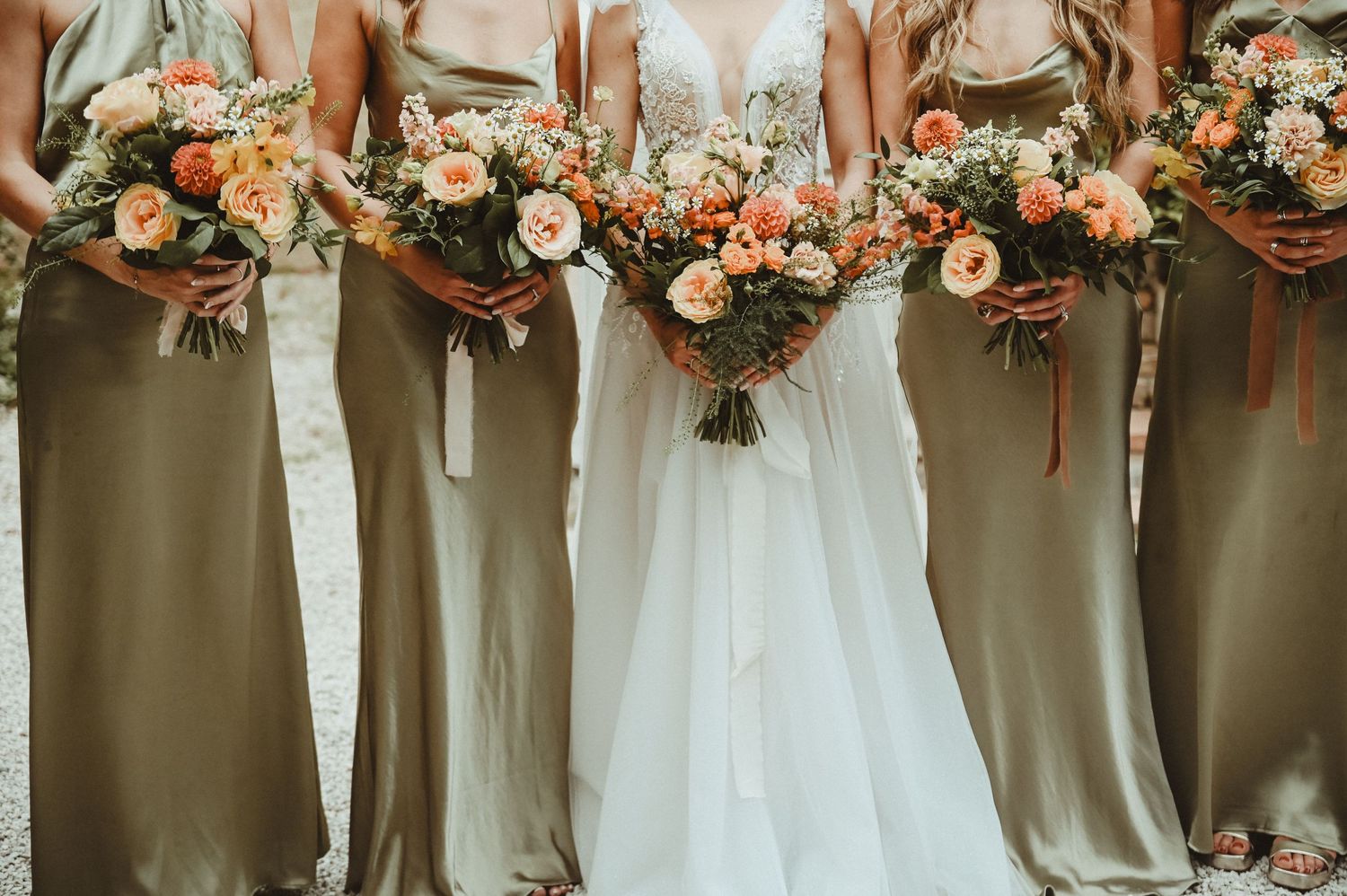 Row of bridesmaids holding peach and coral bouquets while wearing sage green dresses.