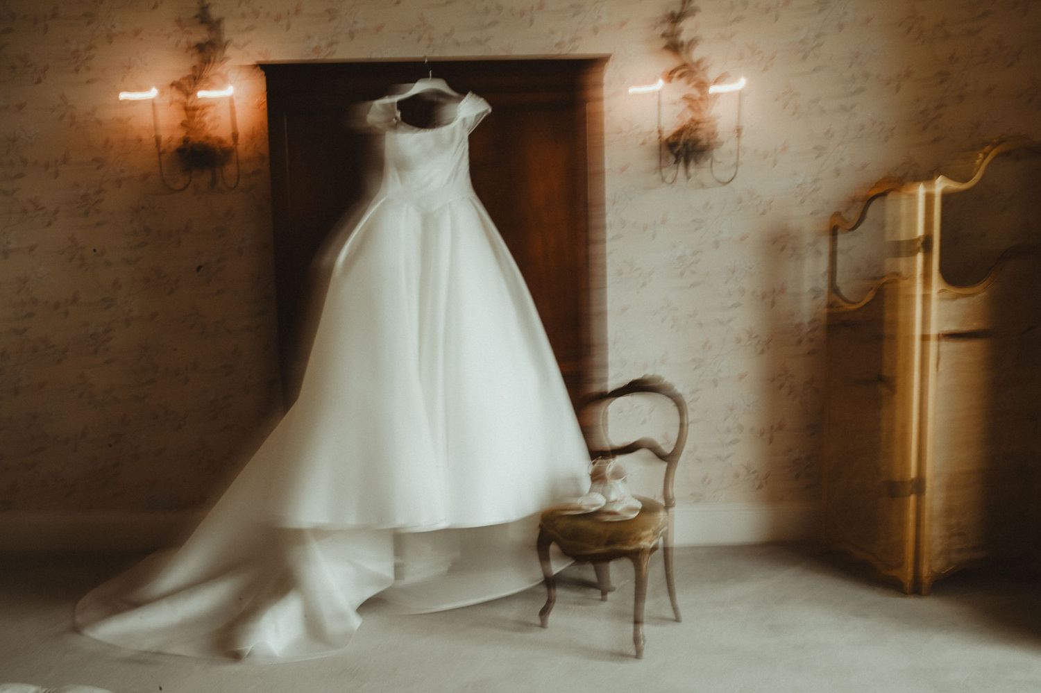 Elegant white wedding dress displayed on a vintage chair against a brick wall with sconces.
