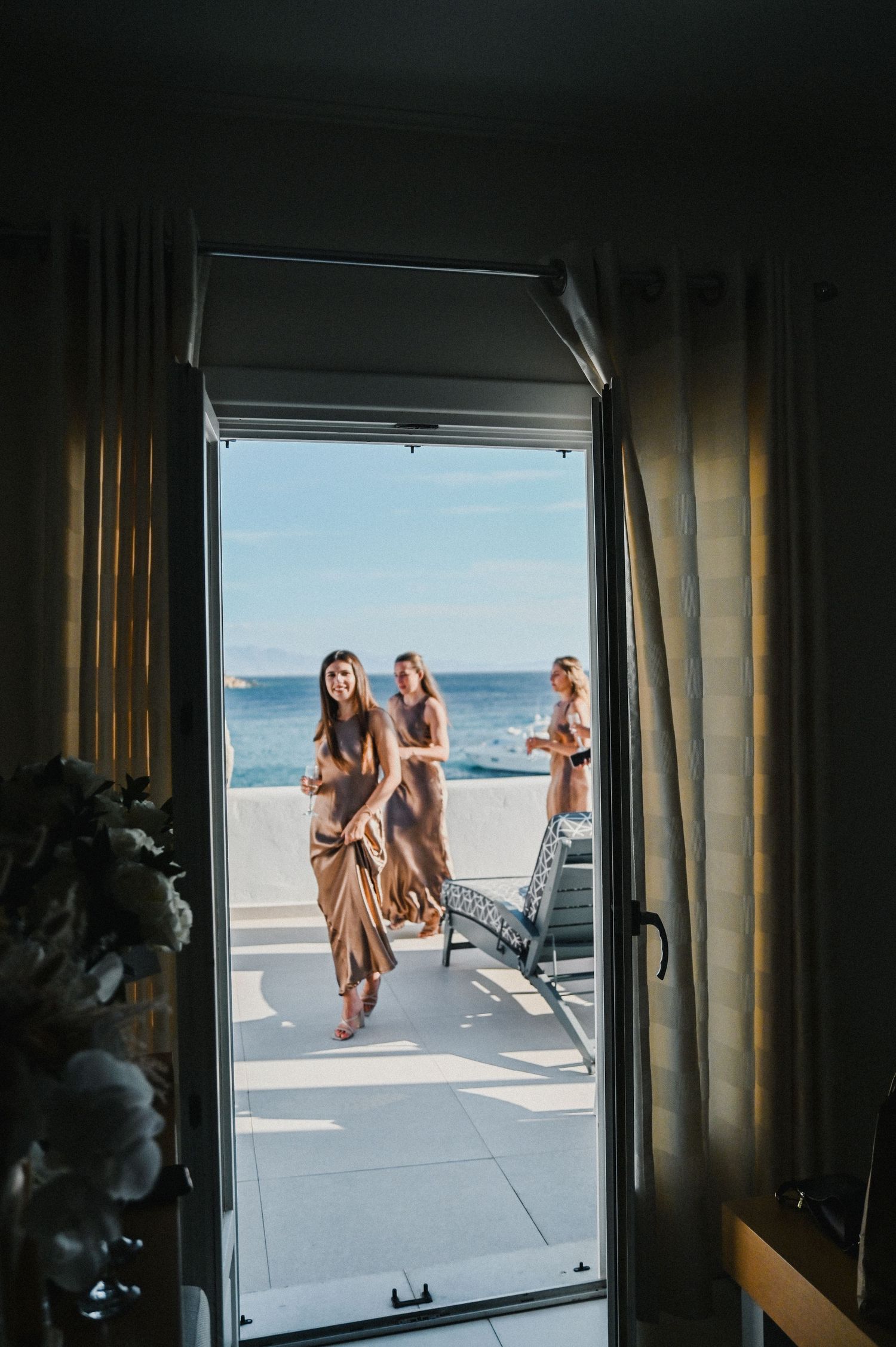 Through an open doorway, guests in elegant dresses pose on a sunlit beachside balcony.