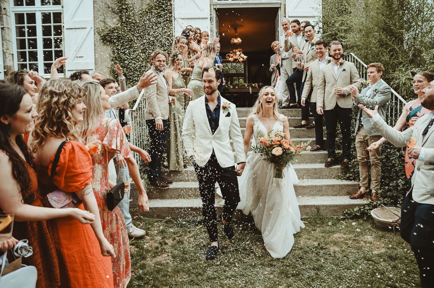 Wedding couple exits historic building while guests throw confetti on stone steps.