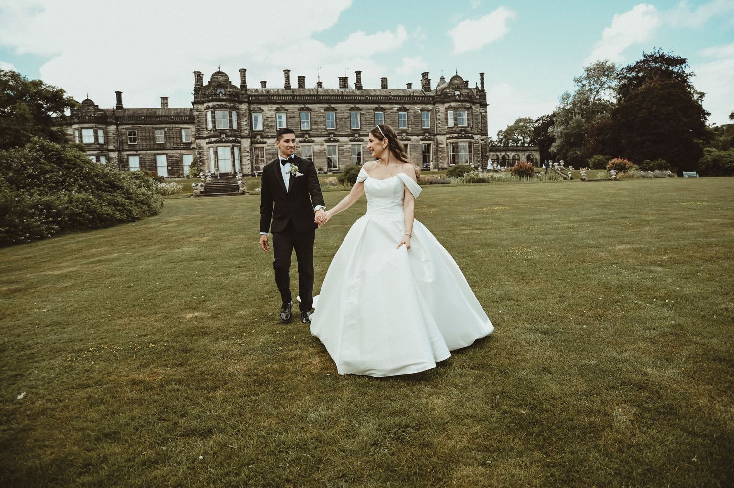 Couple in wedding attire strolls across expansive manor house grounds.