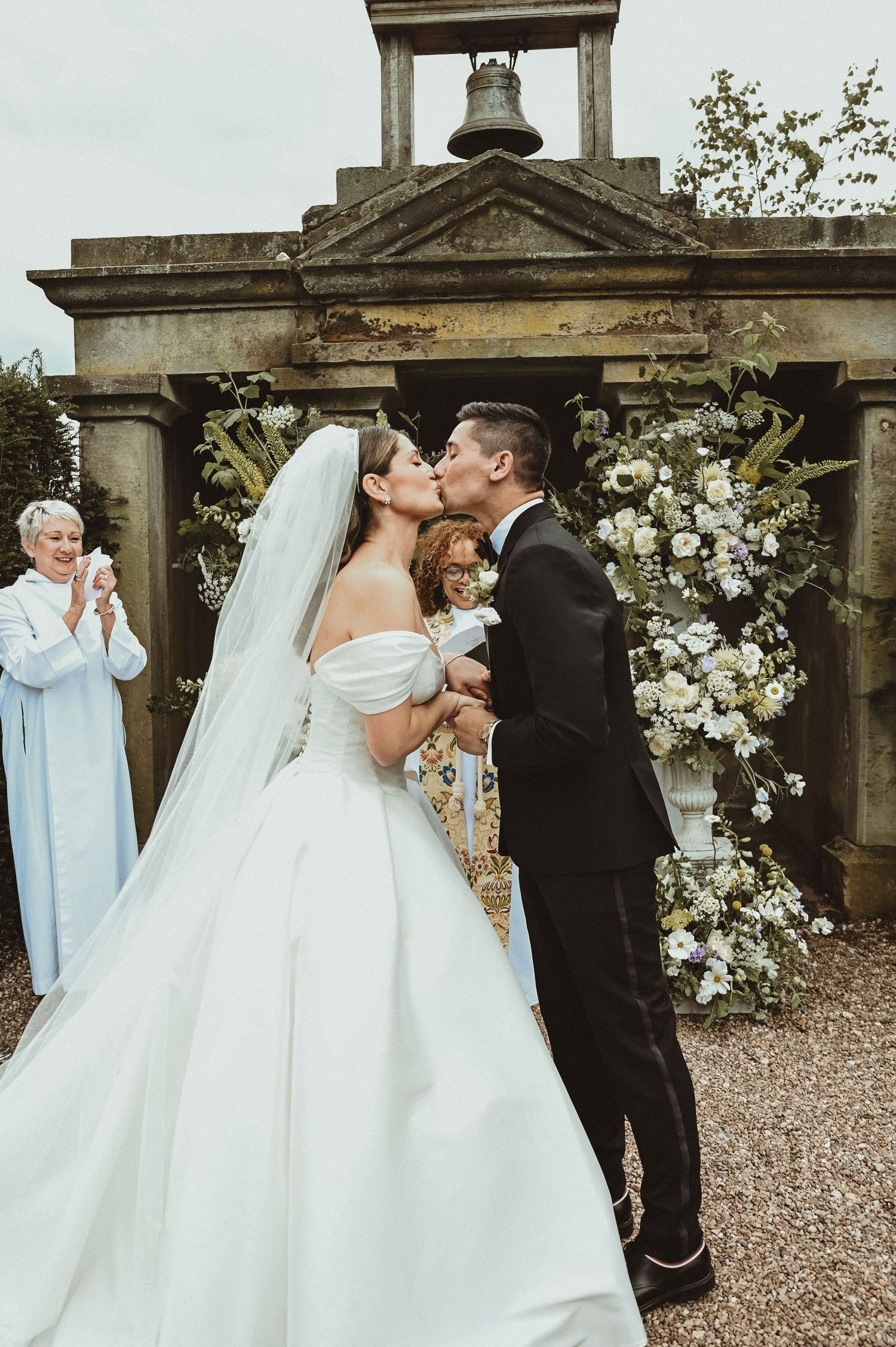 Romantic wedding kiss under an ornate stone bell tower decorated with white floral arrangements.