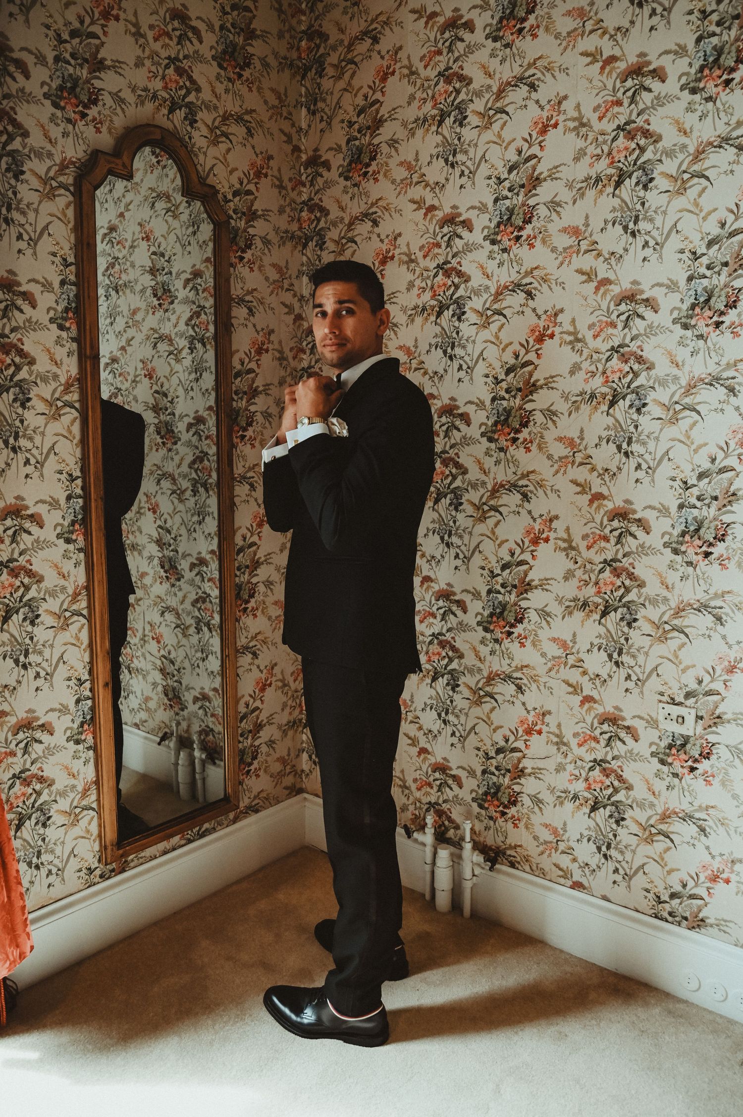 A formal suited figure standing next to an ornate mirror against floral wallpaper.