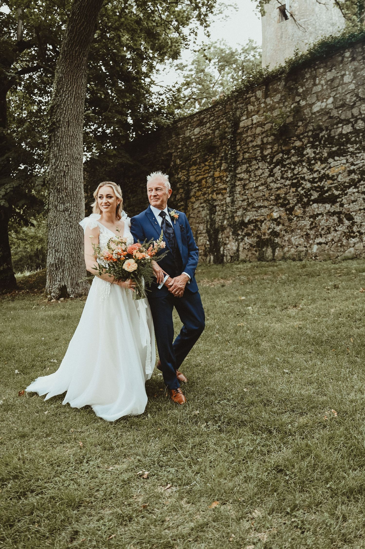 Newlyweds pose together against a stone wall, bride holding a vibrant autumn bouquet.