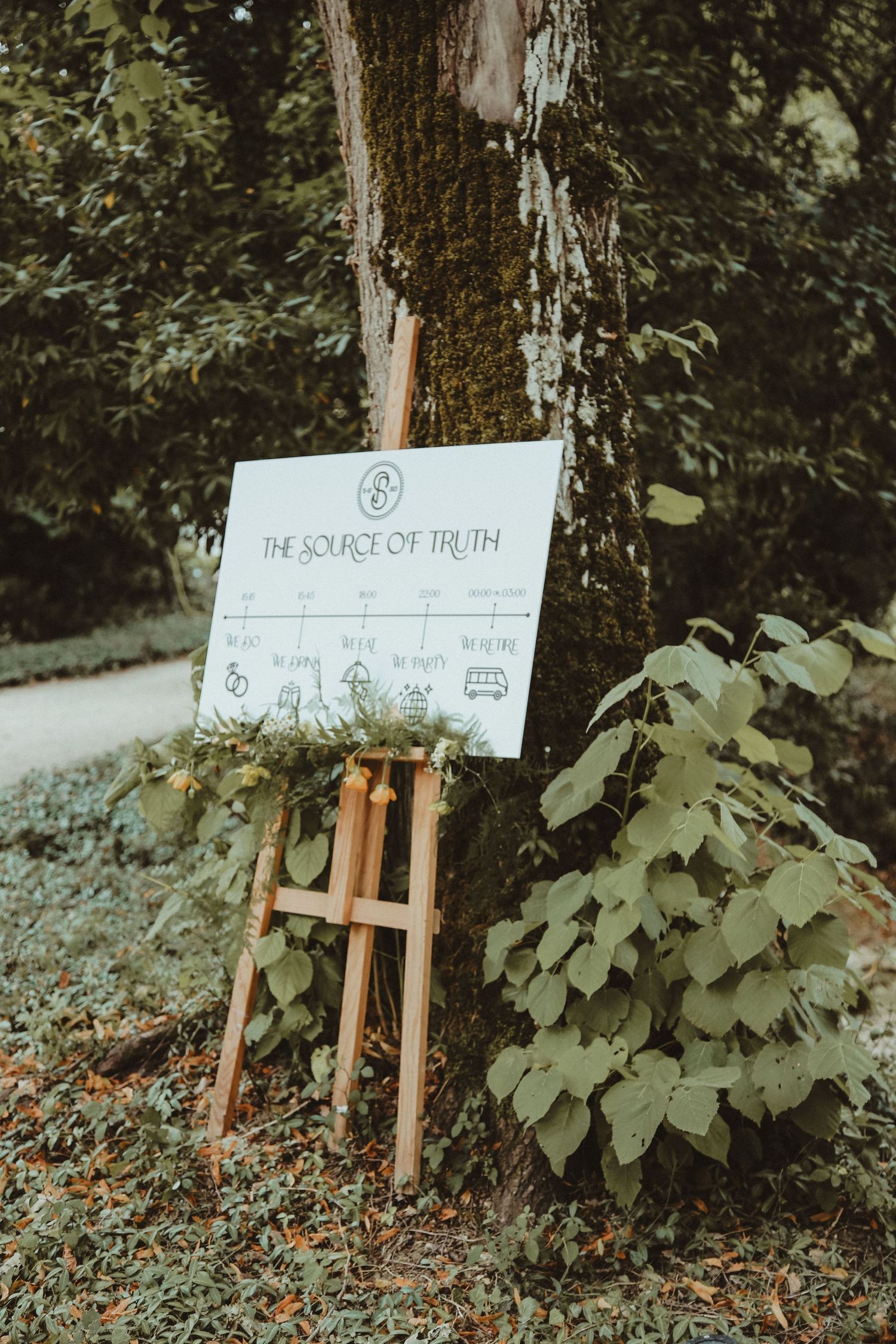 Wedding signage on wooden easel displayed against tree trunk in garden setting.