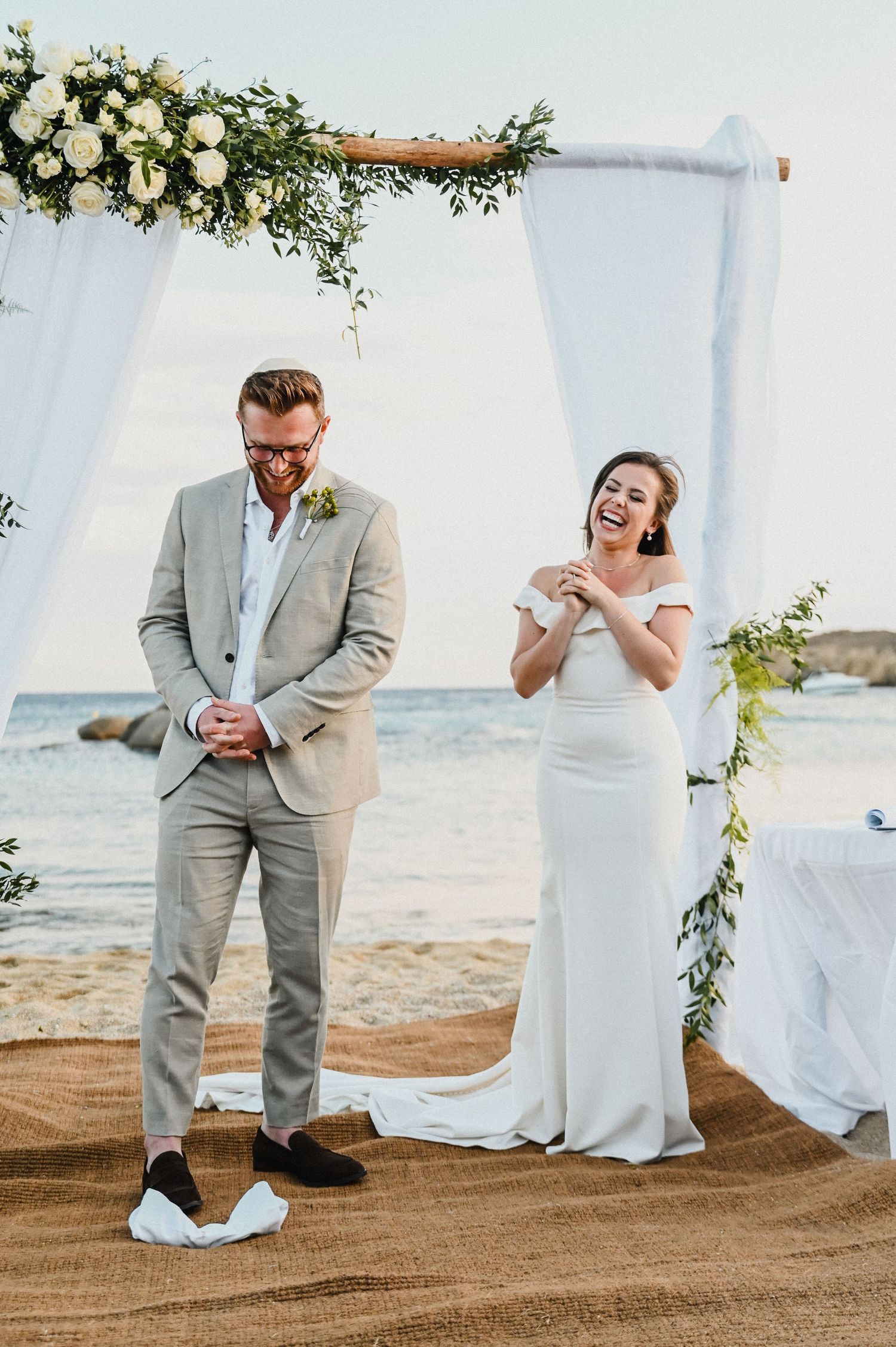 Wedding couple laughing together under flower-adorned arch by the ocean.