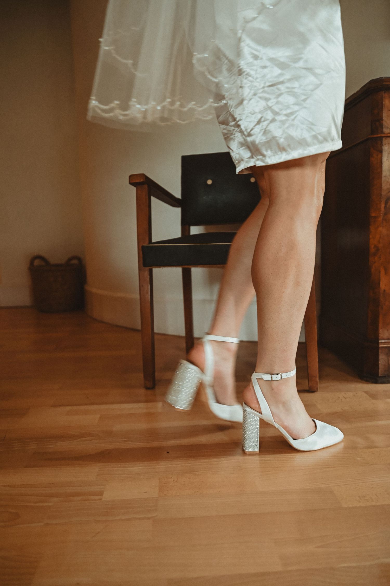 Close-up of white wedding shoes and dress hem on hardwood floor beside wooden chair.