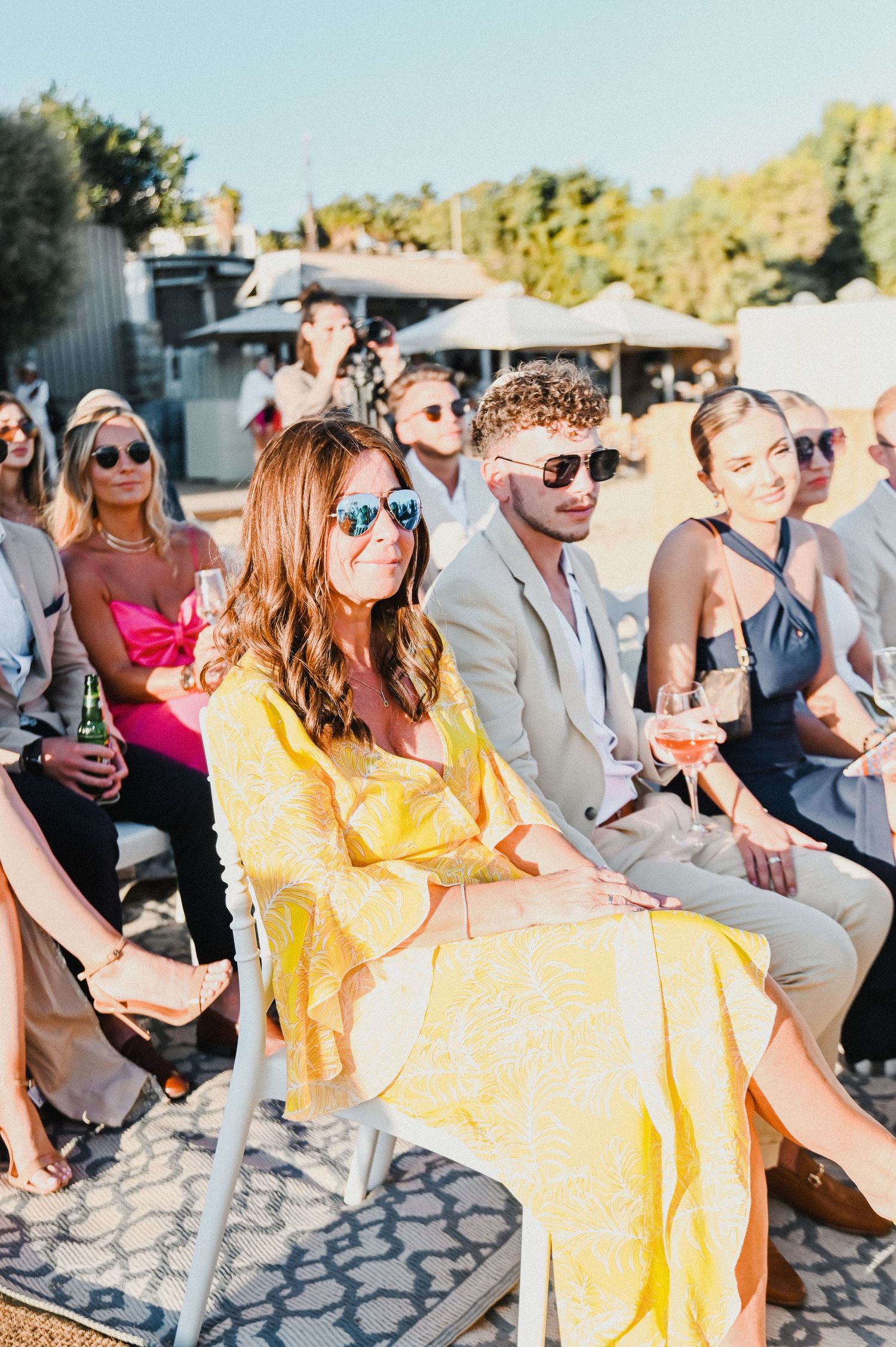 Wedding guests in colorful attire enjoying outdoor beach ceremony with sunglasses.