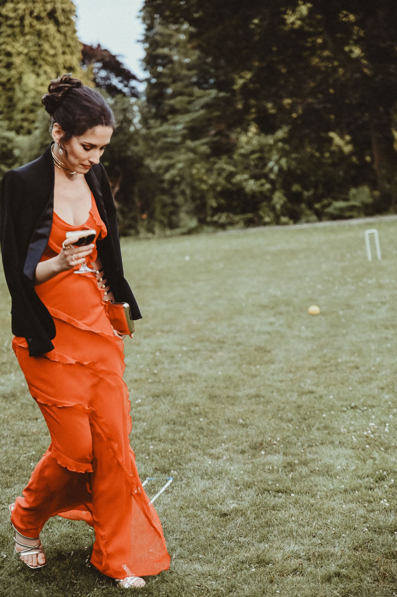 A woman in a vibrant orange dress walks across a lawn with croquet equipment visible in the background.