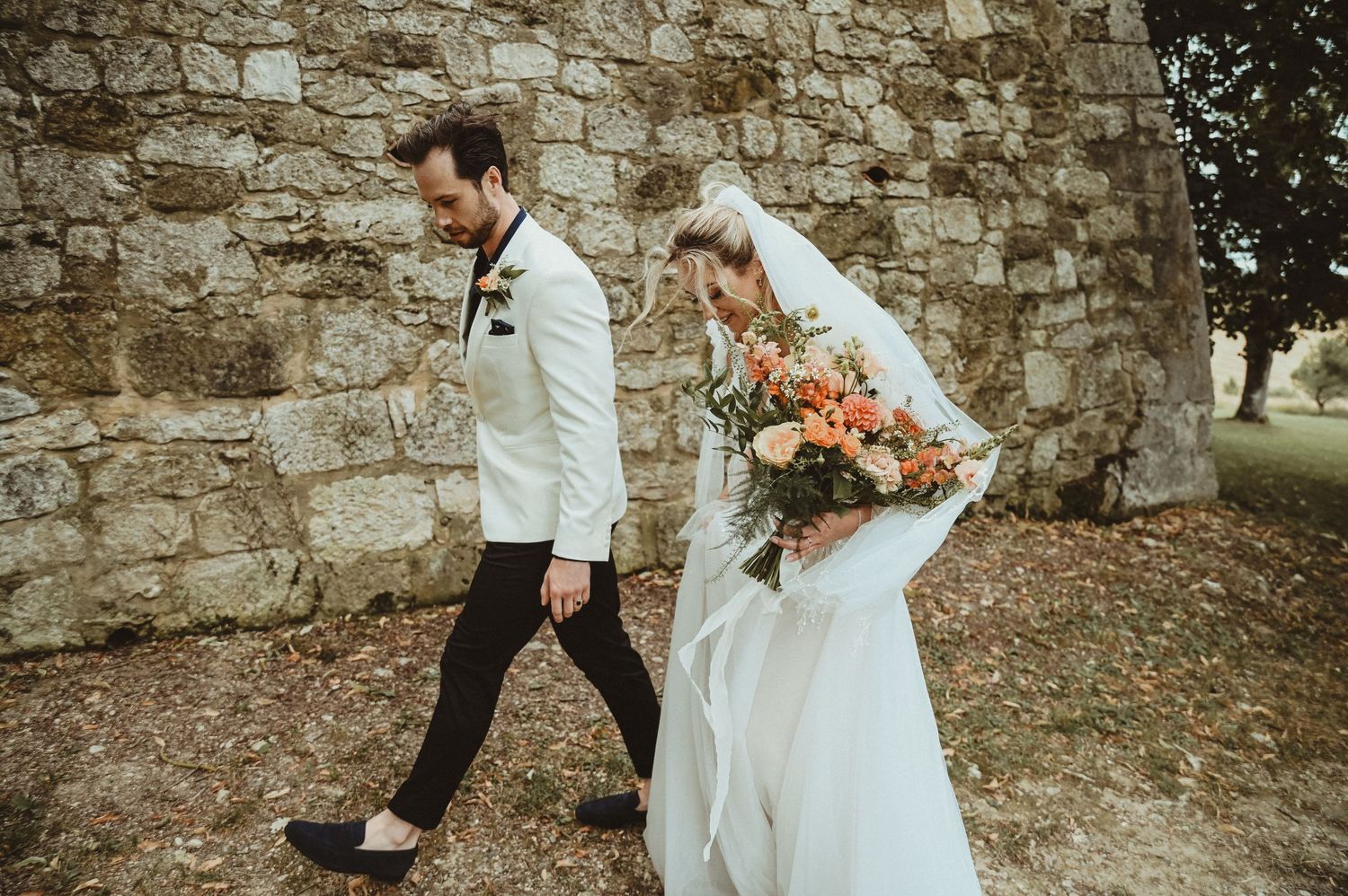 A wedding couple walks along a rustic stone wall with the bride carrying a coral-colored bouquet.