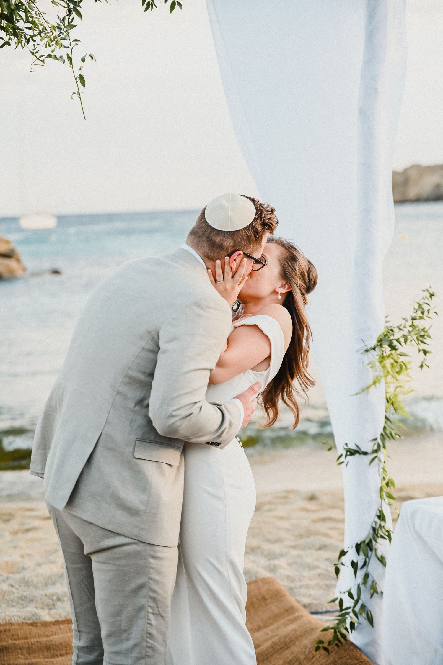 Newlyweds share intimate moment under white draped arch with ocean view.