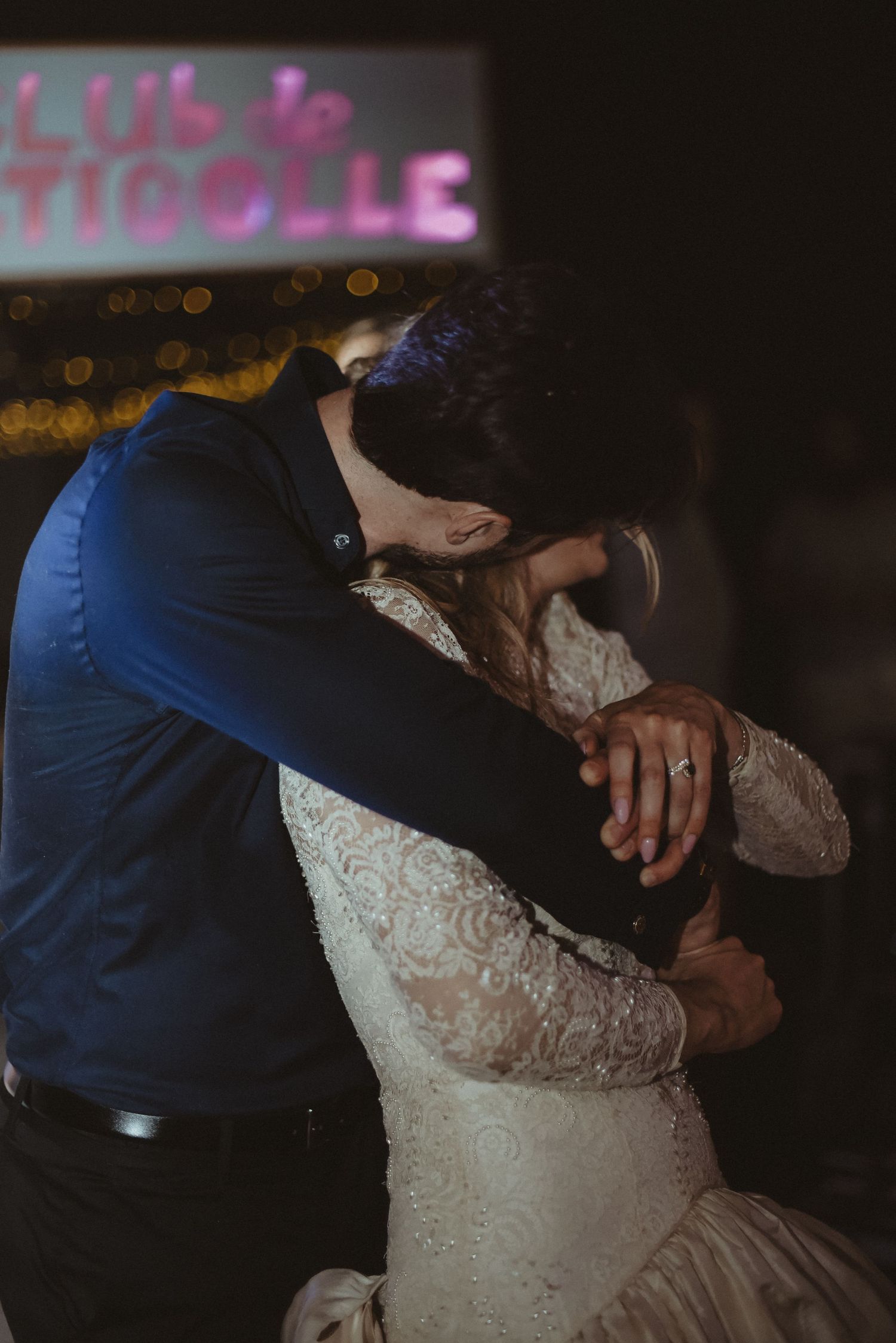 A couple embraces during a dance beneath a neon sign at their celebration.