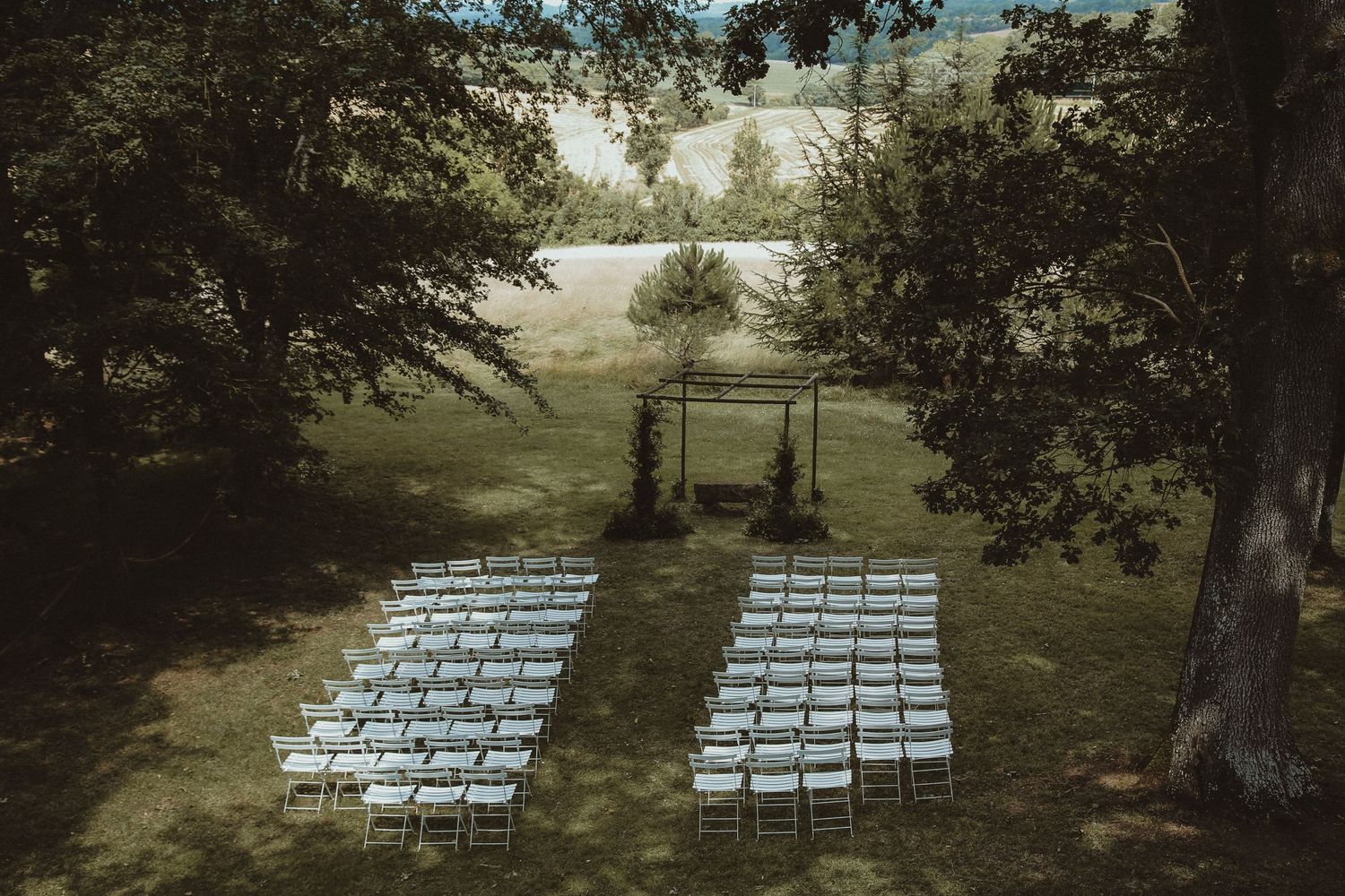 Wedding ceremony setup with white chairs arranged in rows under trees with wooden arch.