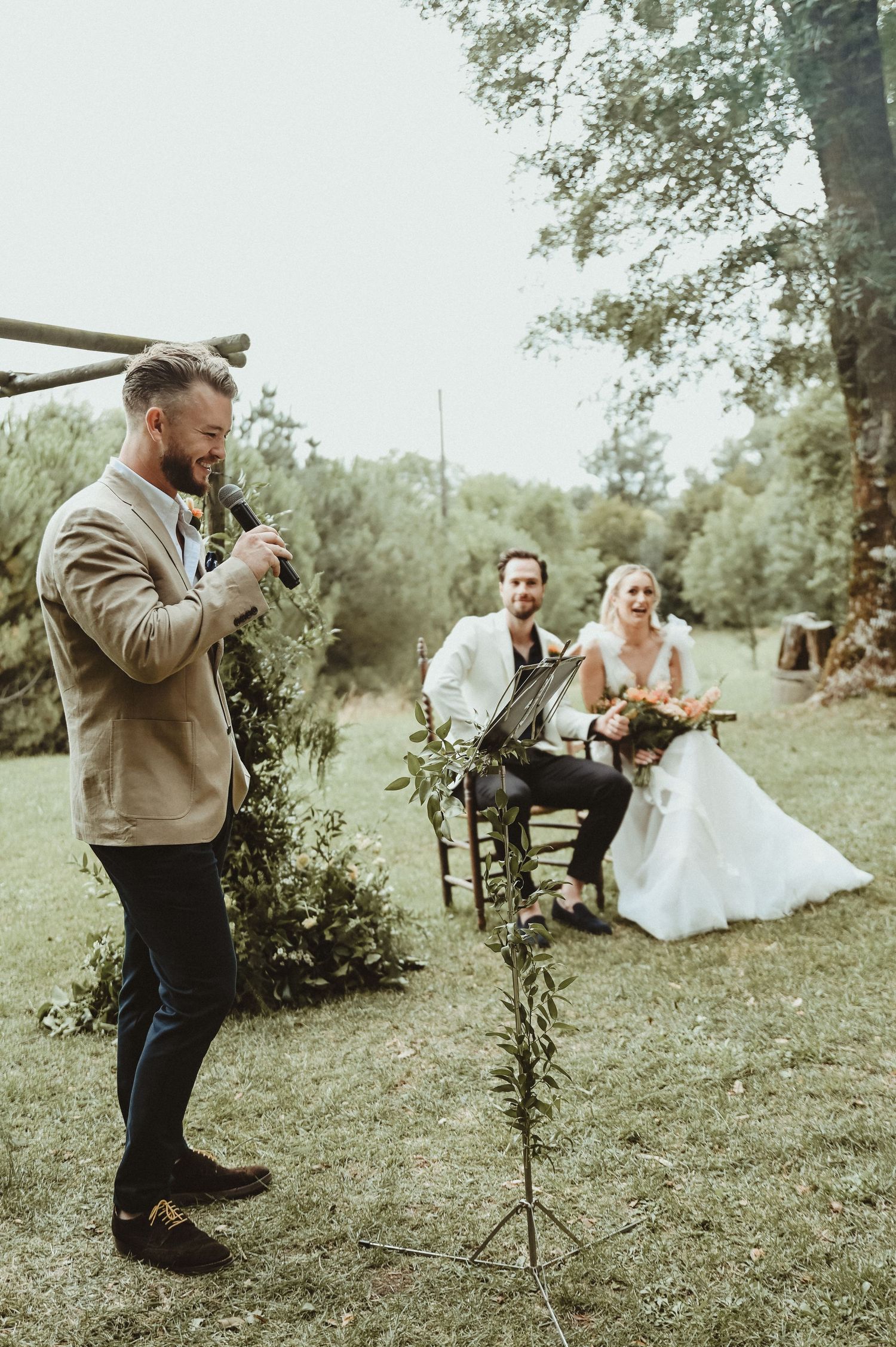 A speaker delivers a speech at an outdoor wedding while the newlyweds listen from decorative chairs.