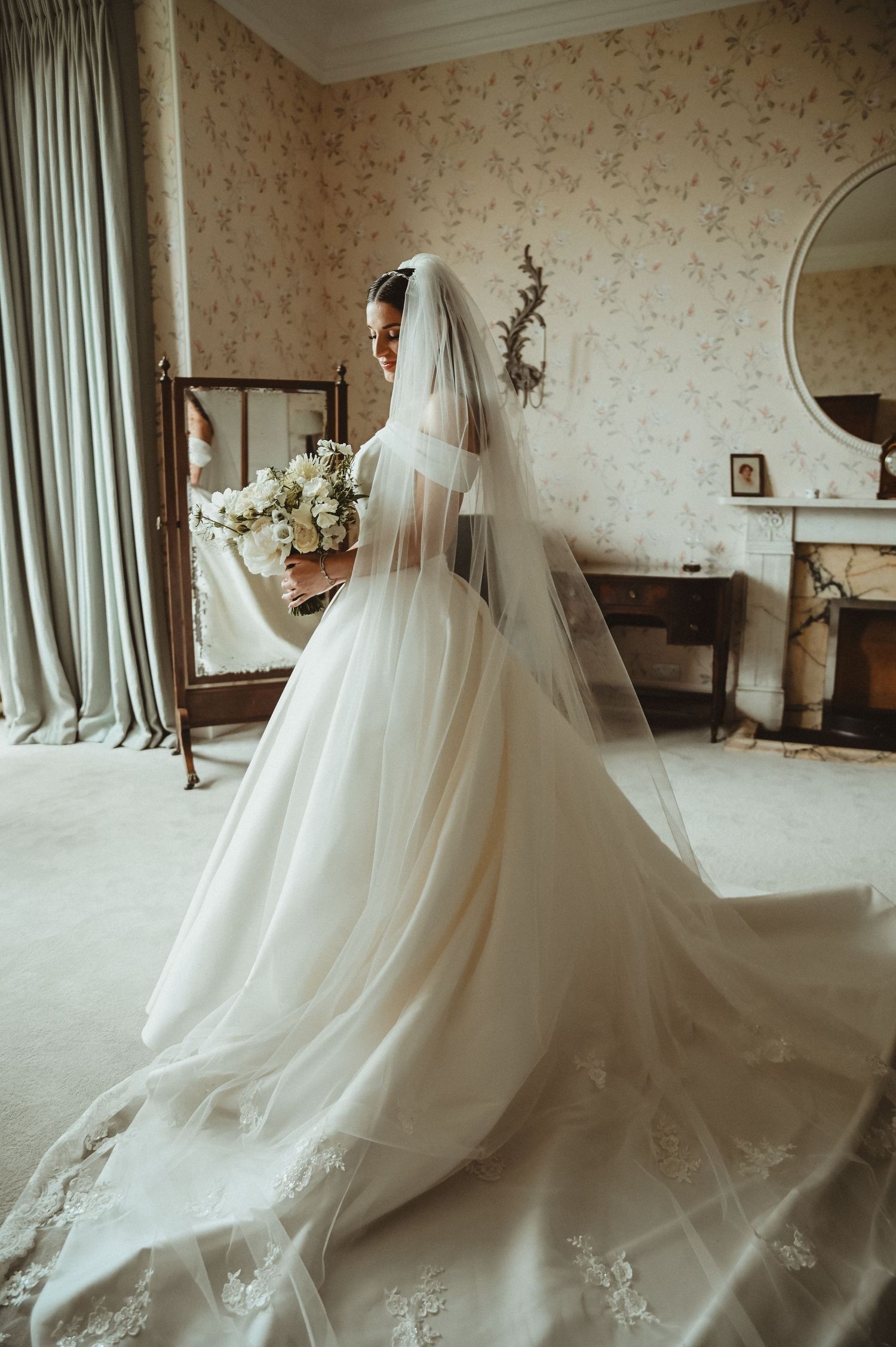 A flowing wedding gown and veil captured in front of an antique mirror in warm, natural light.