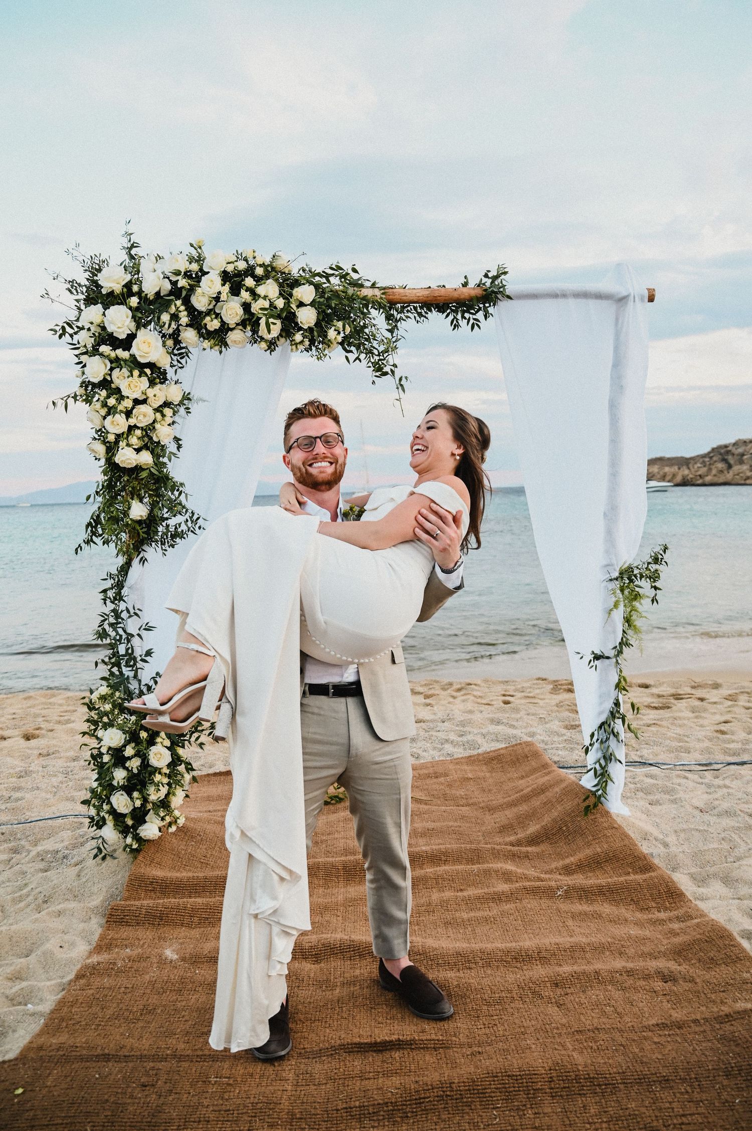 A joyful moment under a floral wedding arch as a couple celebrates on a Mediterranean beach.