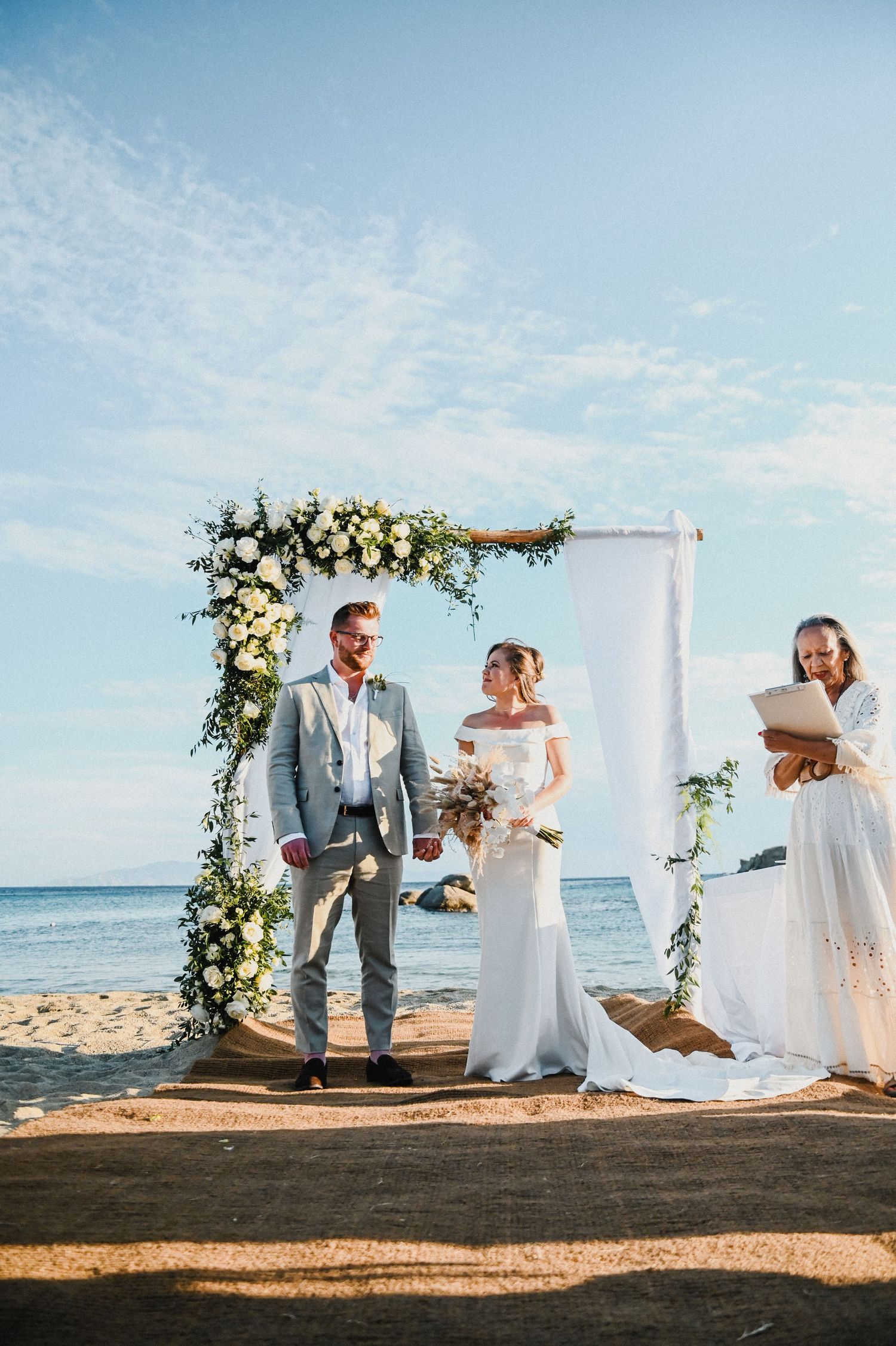 Wedding ceremony on beach with floral arch and couple exchanging vows by the ocean.