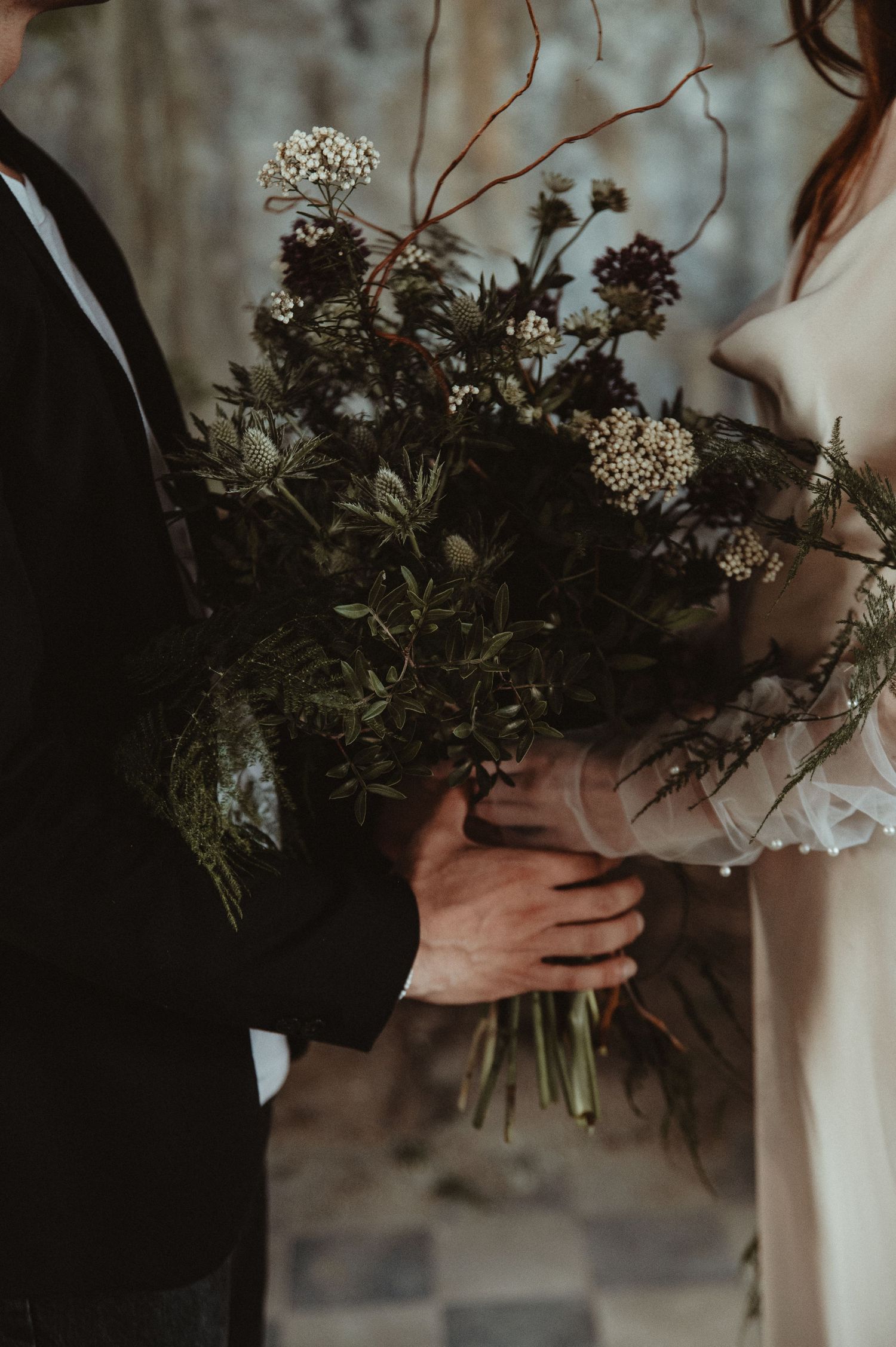 Close-up of a wildflower wedding bouquet being held between two people in formal attire.