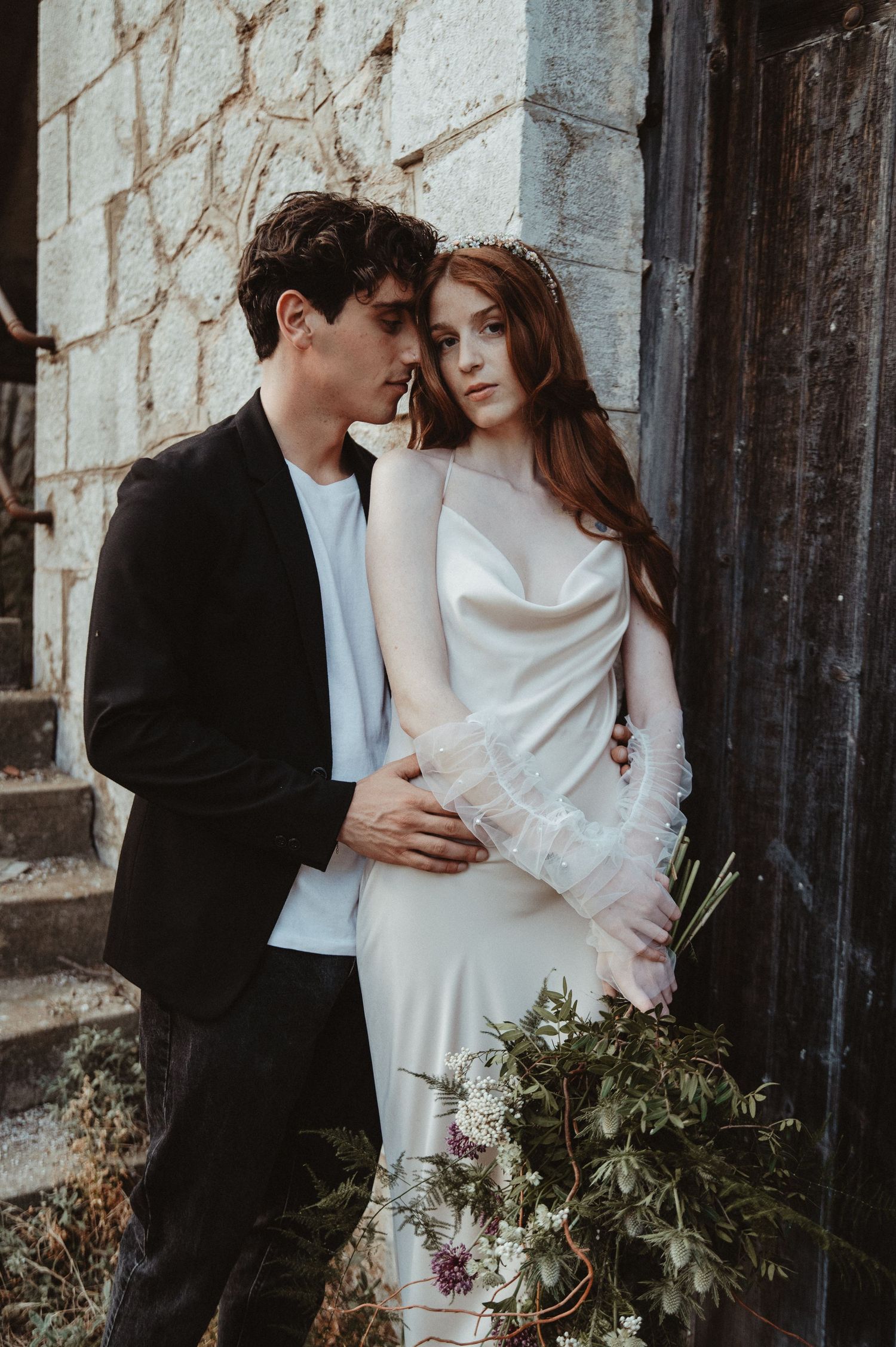 A couple poses on stone steps with a rustic bouquet in elegant wedding attire.