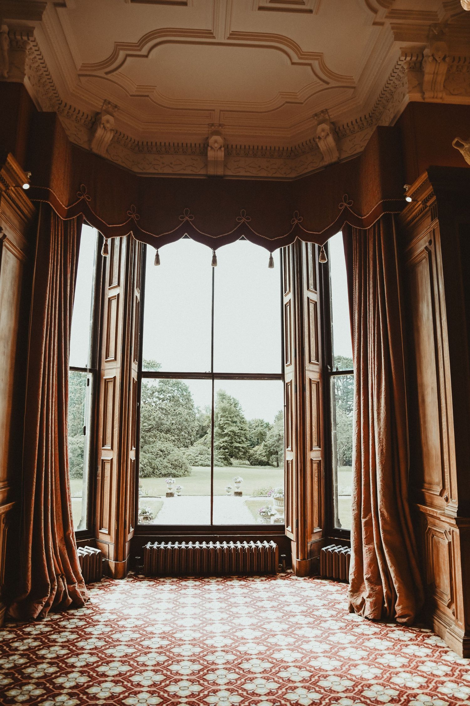 Elegant Victorian bay window with ornate curtains and moldings overlooking a scenic garden view.