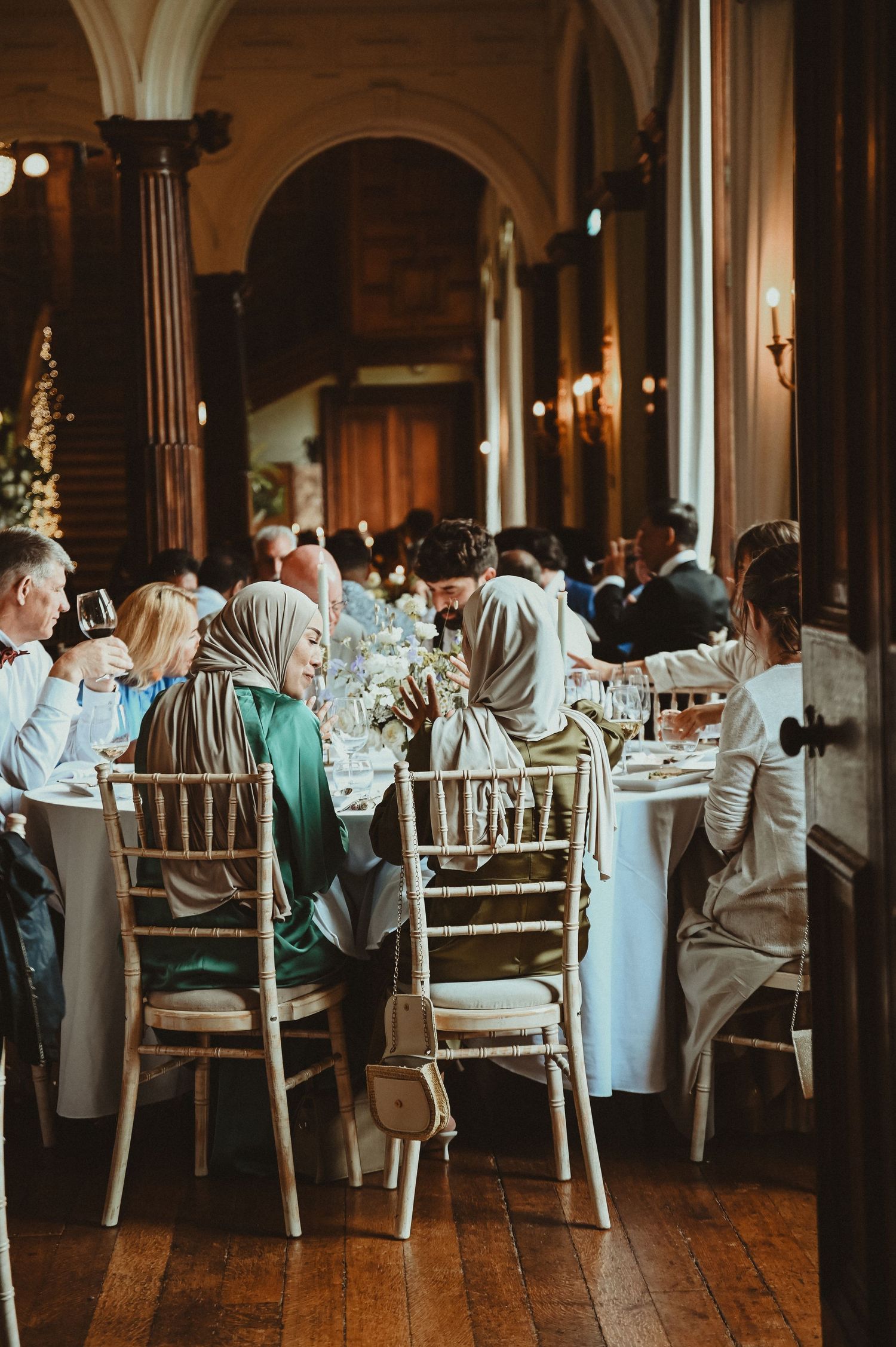 Wedding guests seated at elegant tables in a grand dining hall with arched columns and wooden floors.