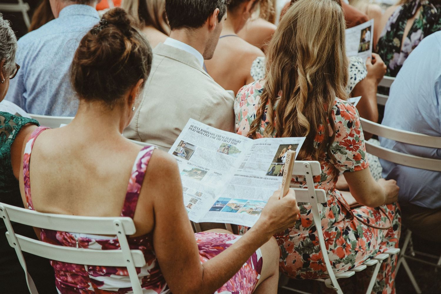 Guests reading wedding programs while seated in white folding chairs at outdoor ceremony.