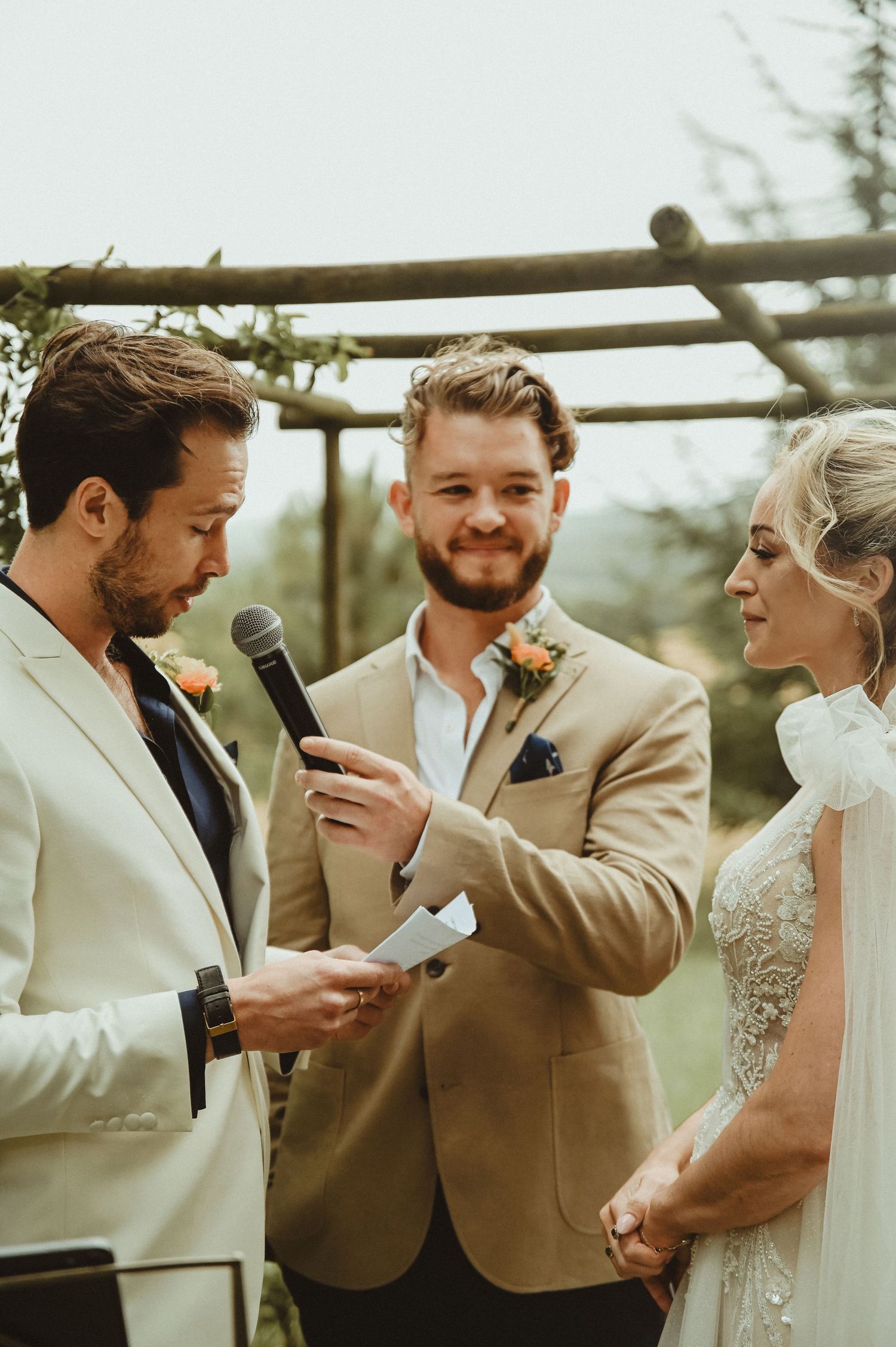 A wedding ceremony underway with an officiant speaking into a microphone beneath a wooden pergola.