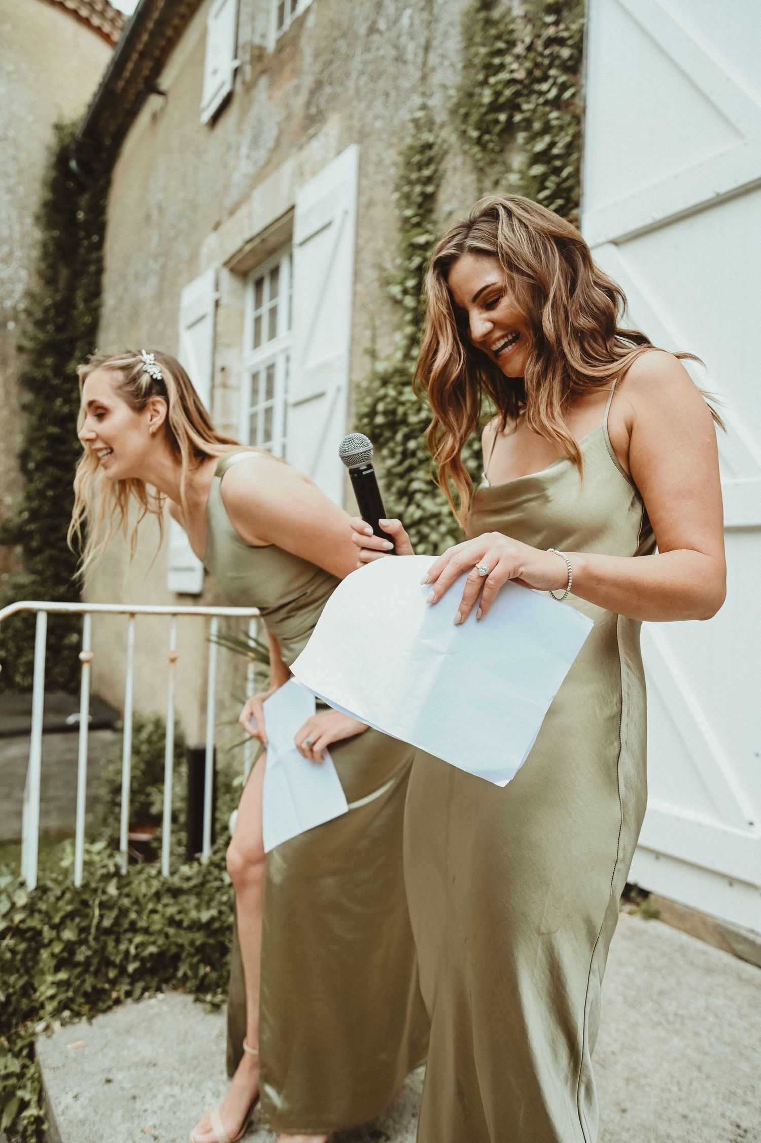 Bridesmaids in olive dresses share a laugh while holding papers during wedding speeches.
