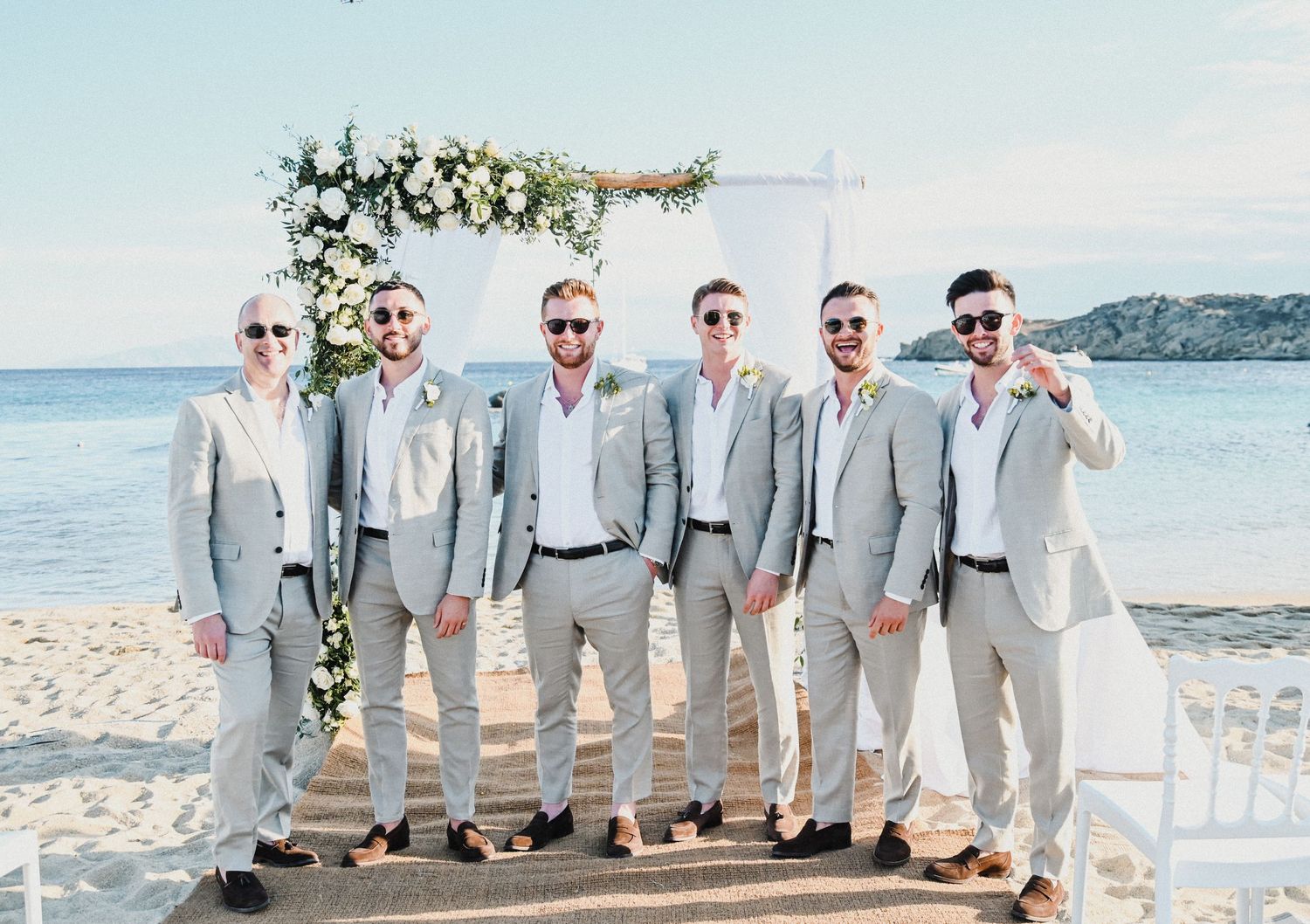 Wedding party in coordinated light gray suits poses on beach with floral arch and ocean view.