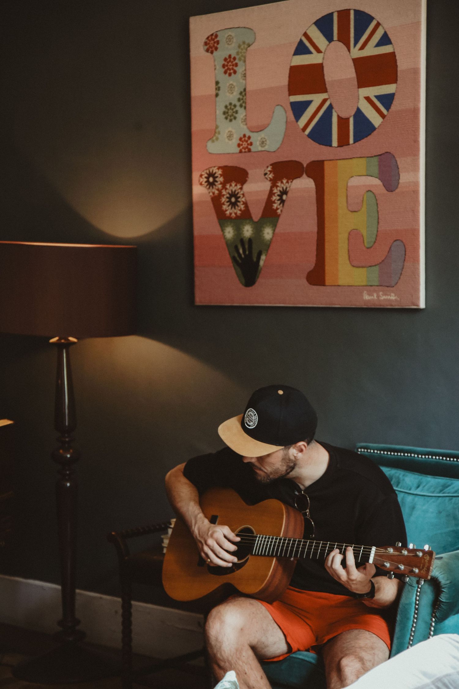 A musician plays acoustic guitar while seated beneath a colorful 'LOVE' wall art featuring a Union Jack design.