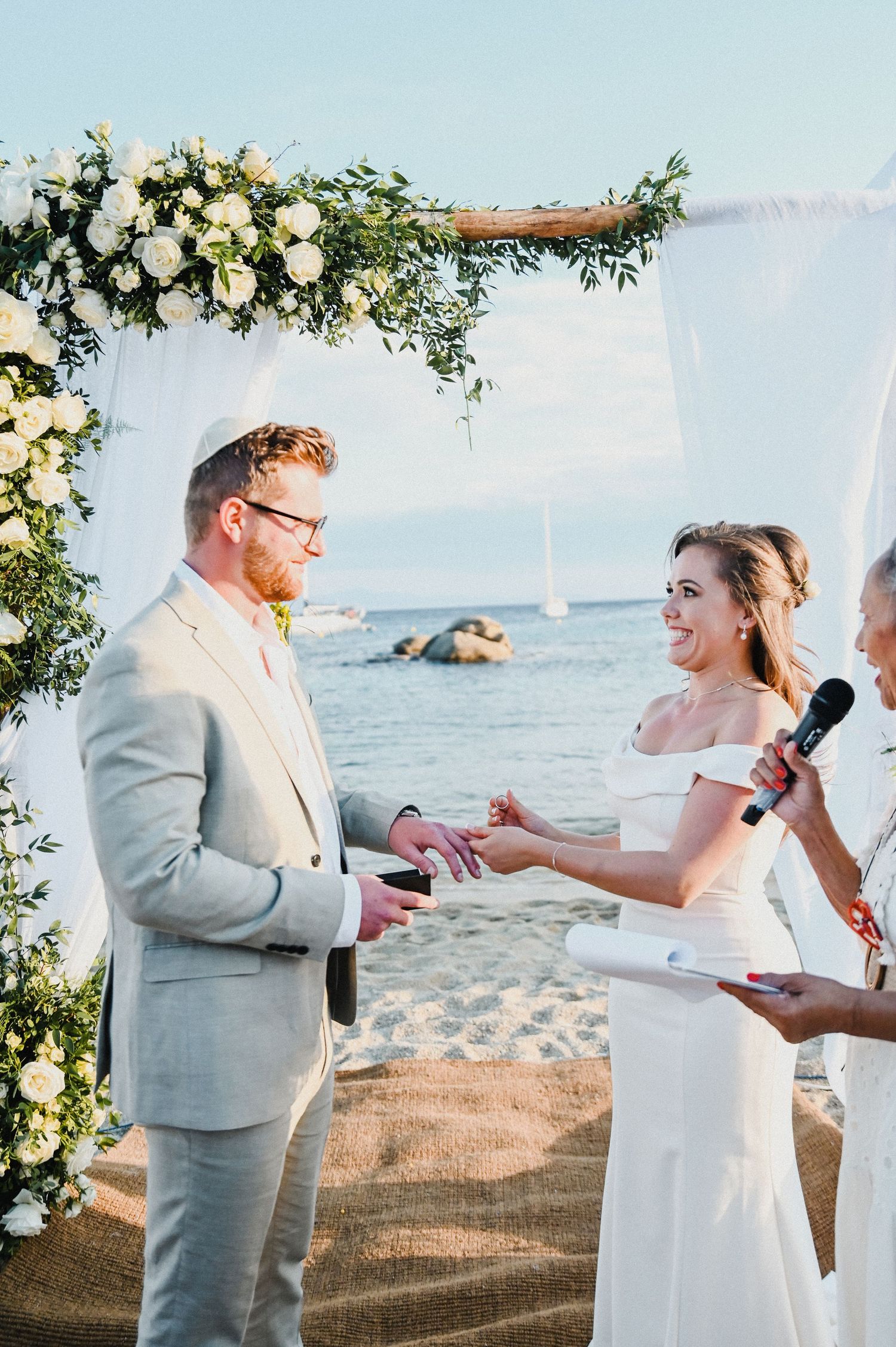 Beachfront wedding ceremony with couple exchanging rings under floral arch.