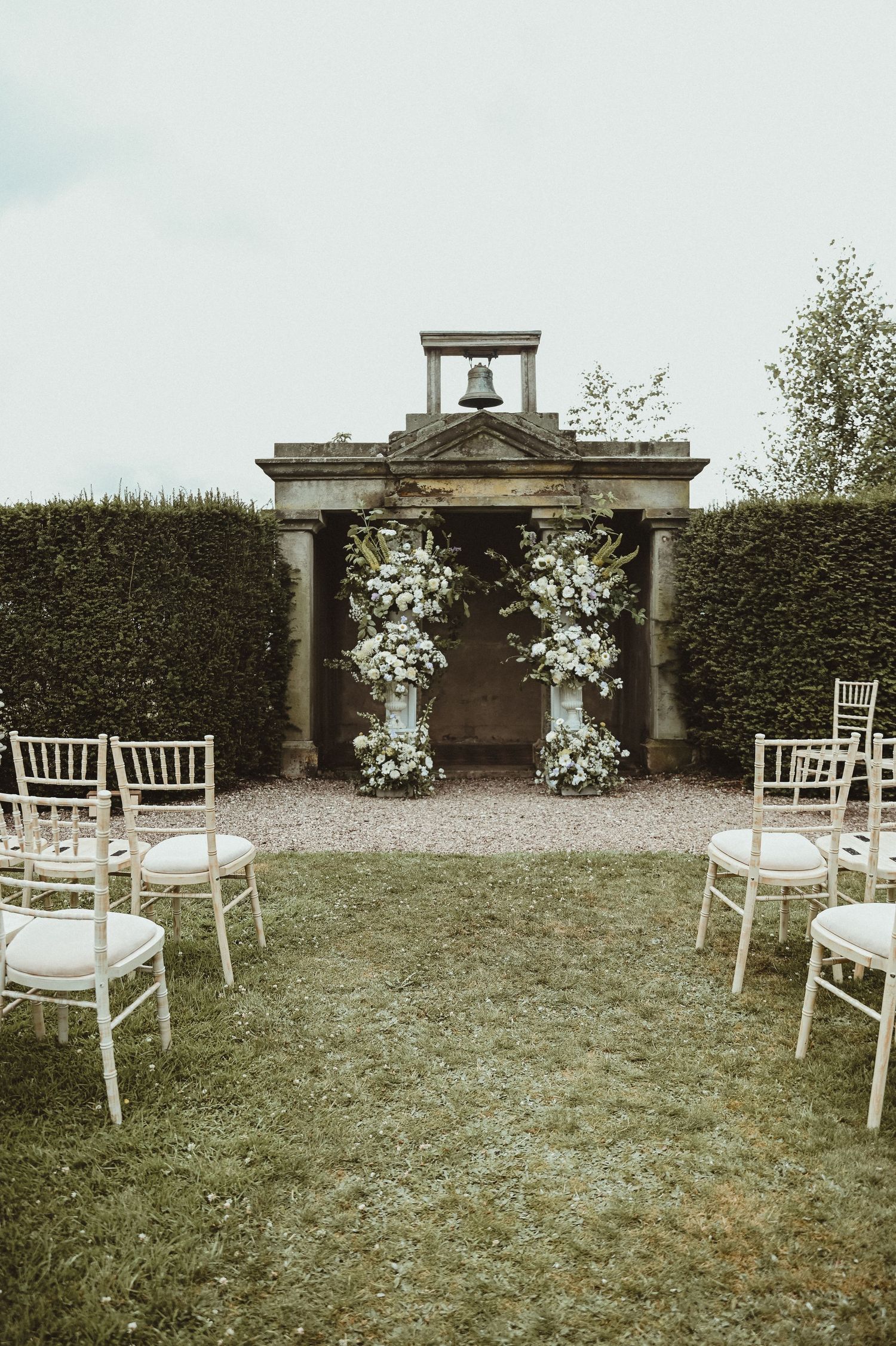 Wedding ceremony setup with white chairs facing a classical stone archway decorated with floral arrangements.