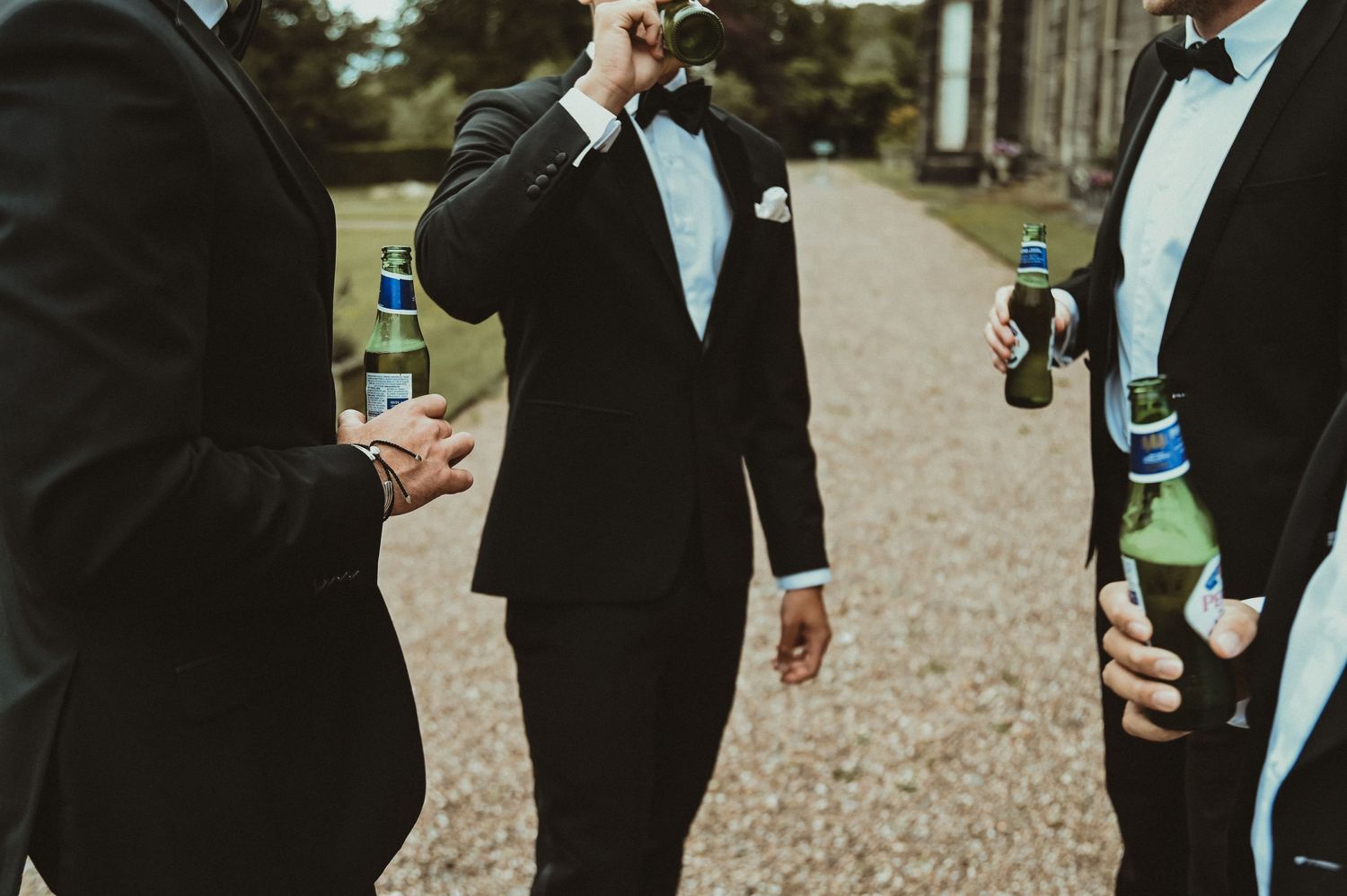 Groomsmen in tuxedos holding beer bottles while socializing outdoors.