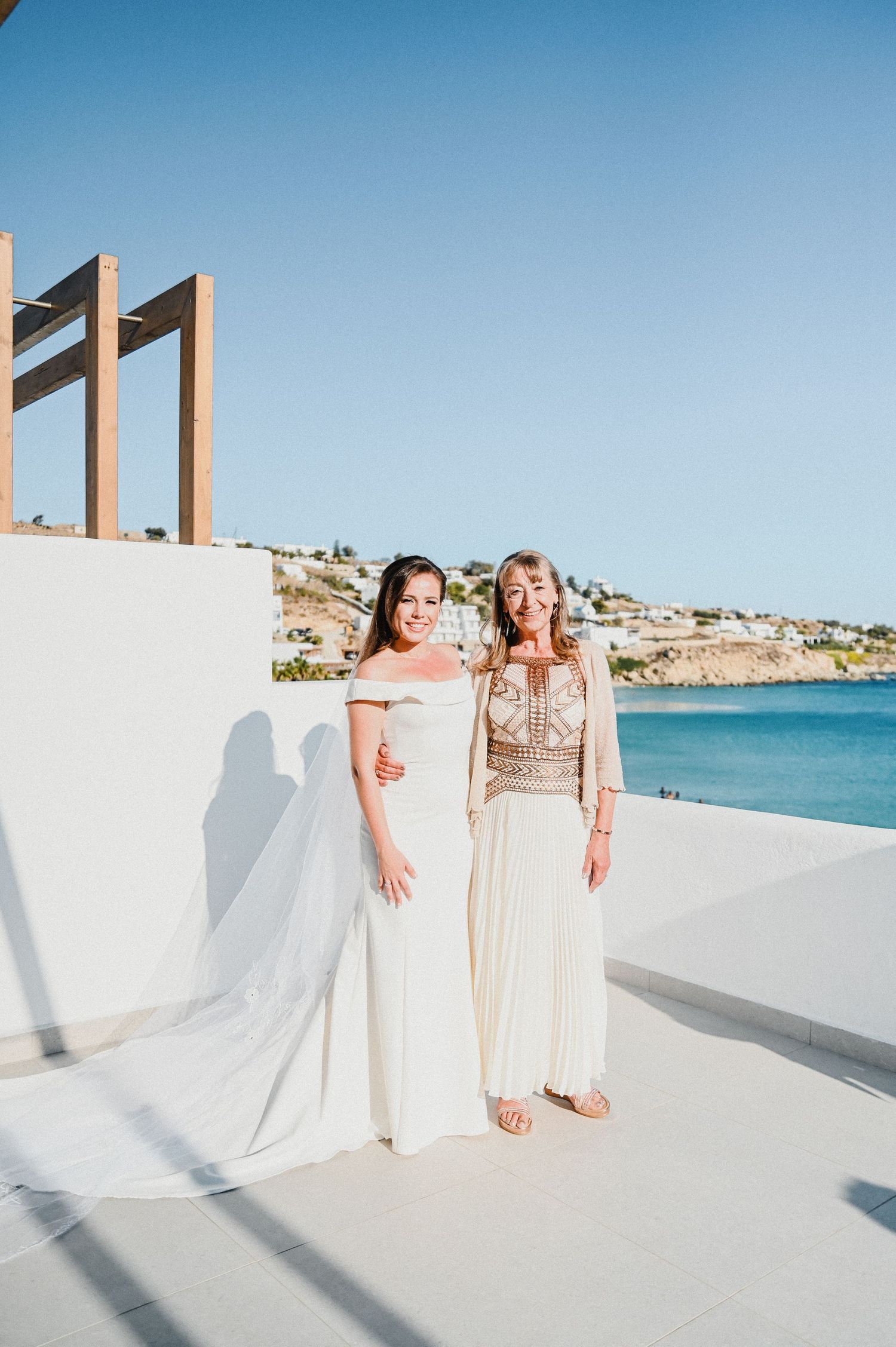Two women in white formal wear stand together on seaside terrace with ocean view.