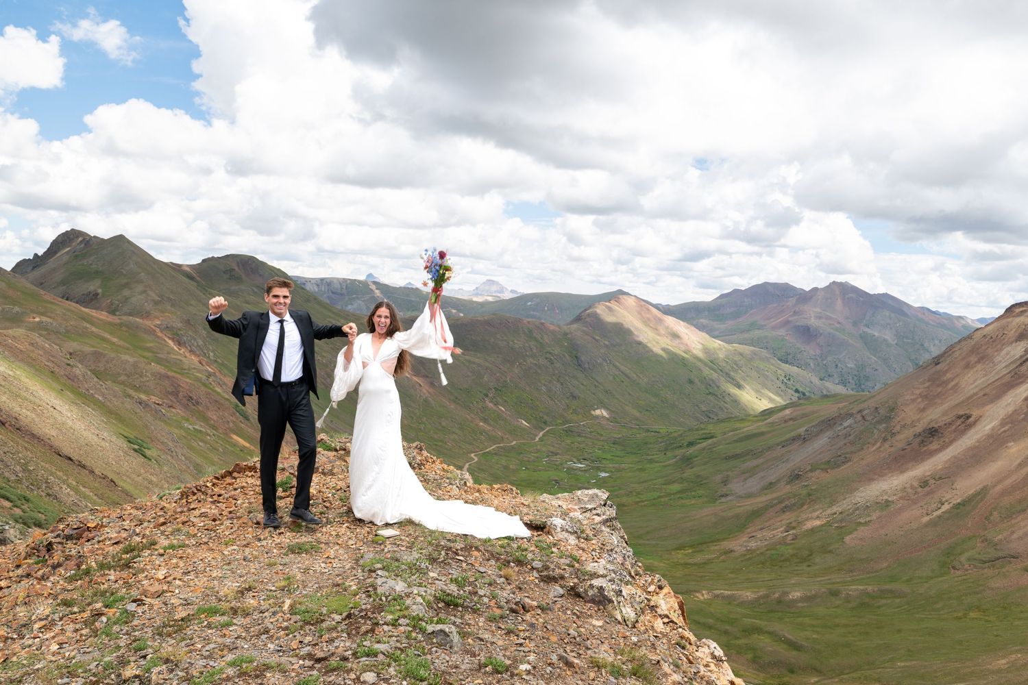 Bride and groom celebrate on mountain peak in wedding attire, surrounded by stunning green valleys and colorful peaks under cloudy sky.