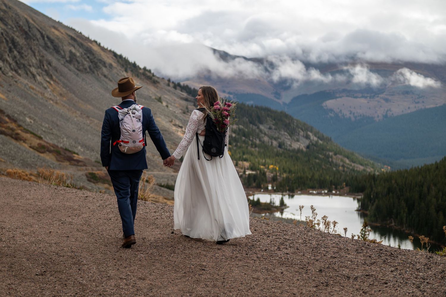 Bride and groom holding hands walking along a mountain trail with a scenic lake and cloudy peaks in the background.