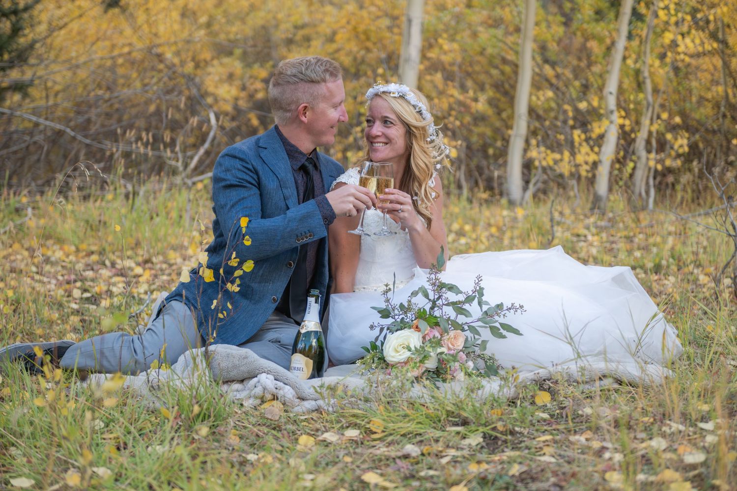 A couple in wedding attire share a champagne toast and cake together on a grassy hill during autumn with fall foliage around them.