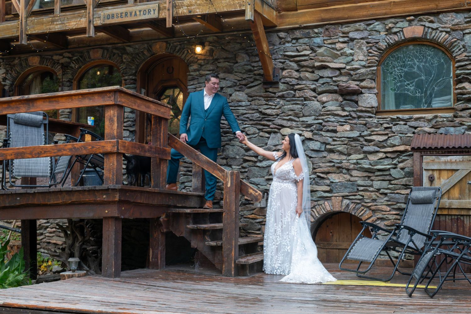 Bride and groom pose on rustic wood and stone  steps, she extends her hand to get  him to help go up the wooden stairs in front of the cabin