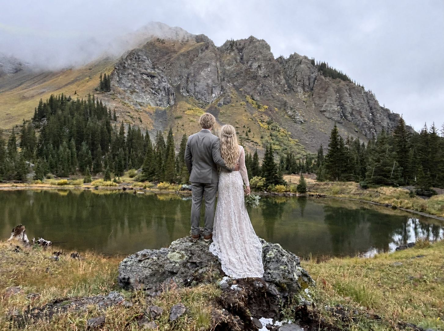 Bride and groom stand on rocky alpine lake shore with misty mountain peaks and evergreen trees backdrop. The grasses are in fall color.