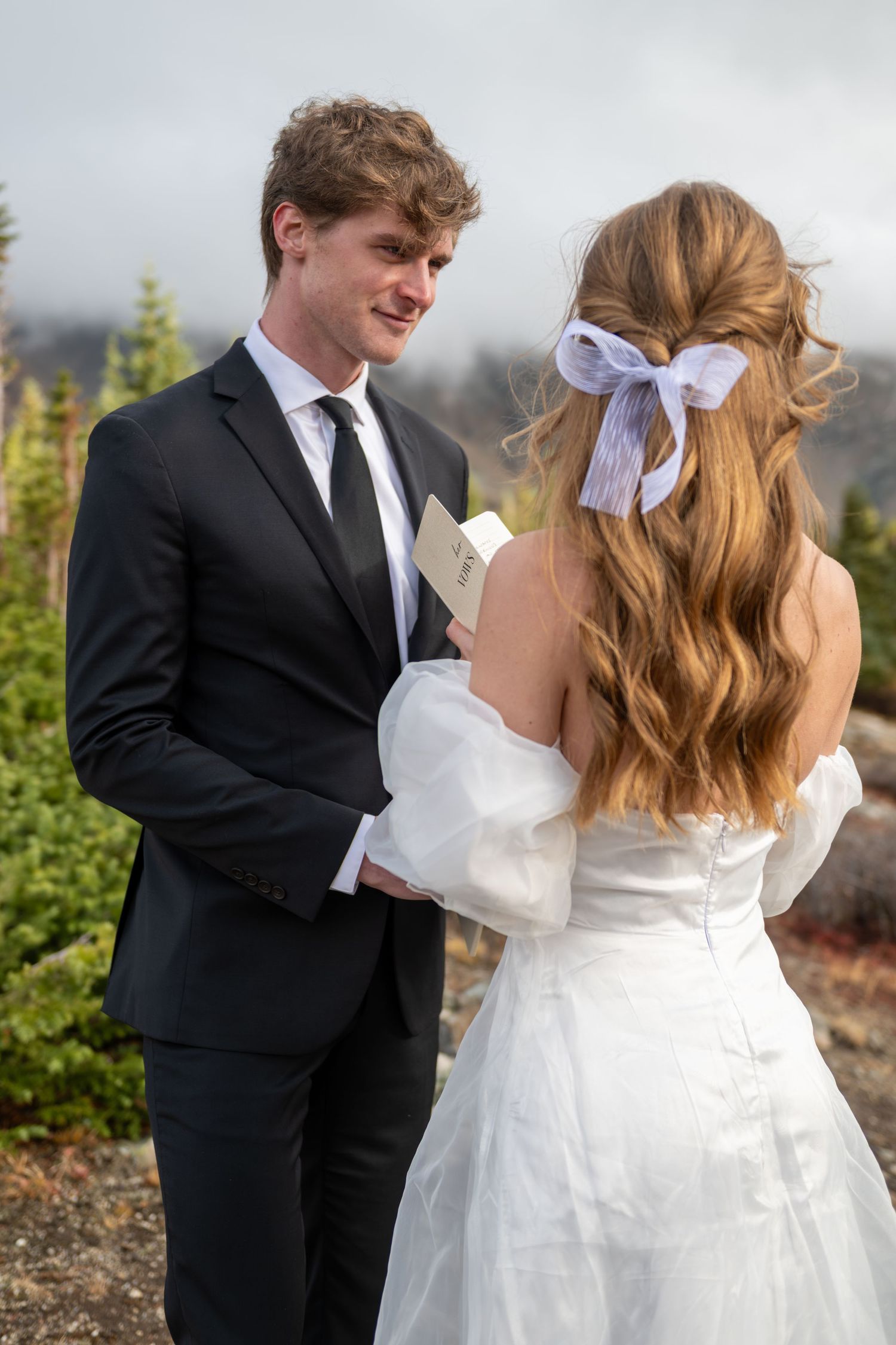 A groom stares lovingly at his bride as she reads her vows during their elopement. Pine trees are behind them and sunset light is present.